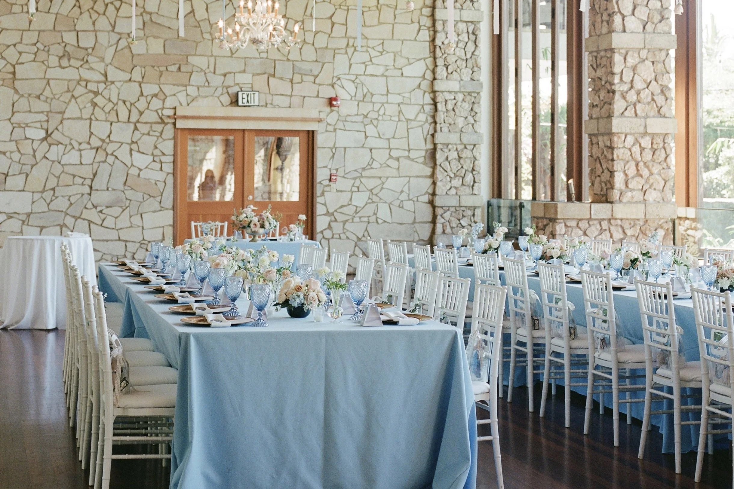 A light-filled indoor wedding reception with blue tablecloths and a stone-paneled wall in the background.