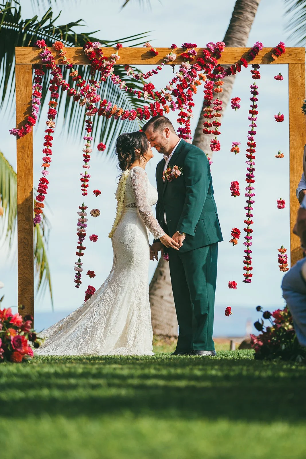 A bride and groom forehead to forehead before their beachside wedding ceremonial arch draped with fuchsia carnations and jewel-tone floral aisle markers.