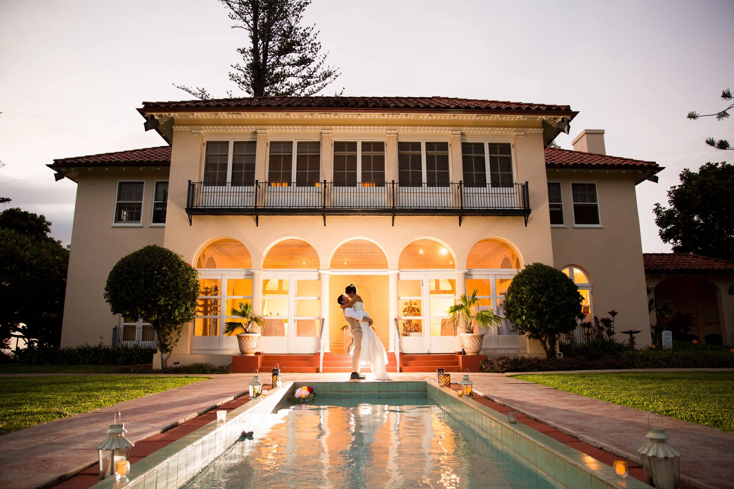 A bride and groom posed between an arch-front manor and a Mediterranean reflection pool