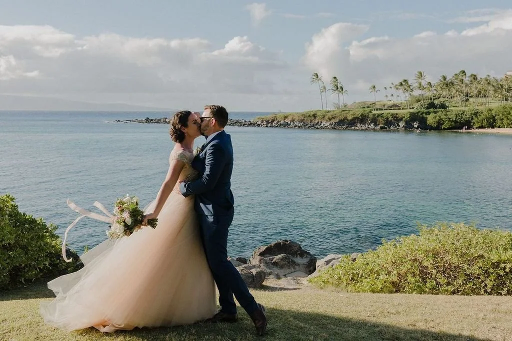 A bride and groom sharing a kiss on the grass before a wide expanse of blue ocean.