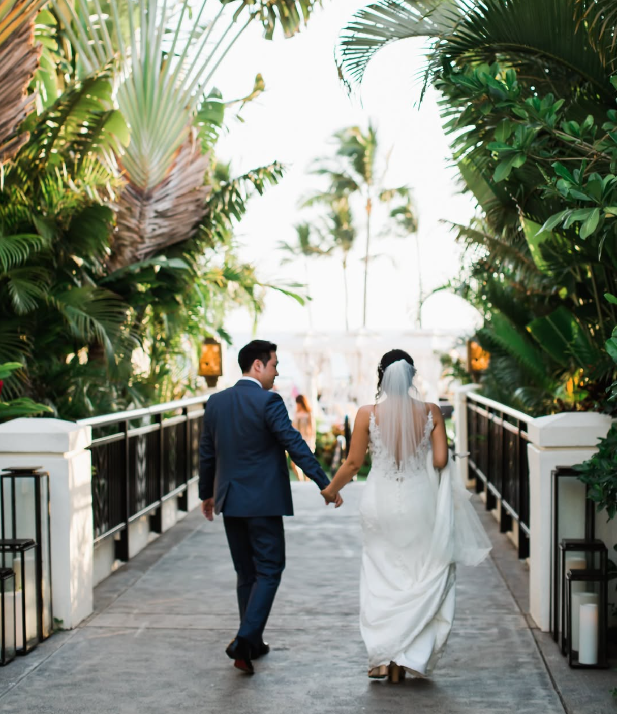 A bride and groom crossing an outdoor hotel bridge, surrounded by dense palm trees.