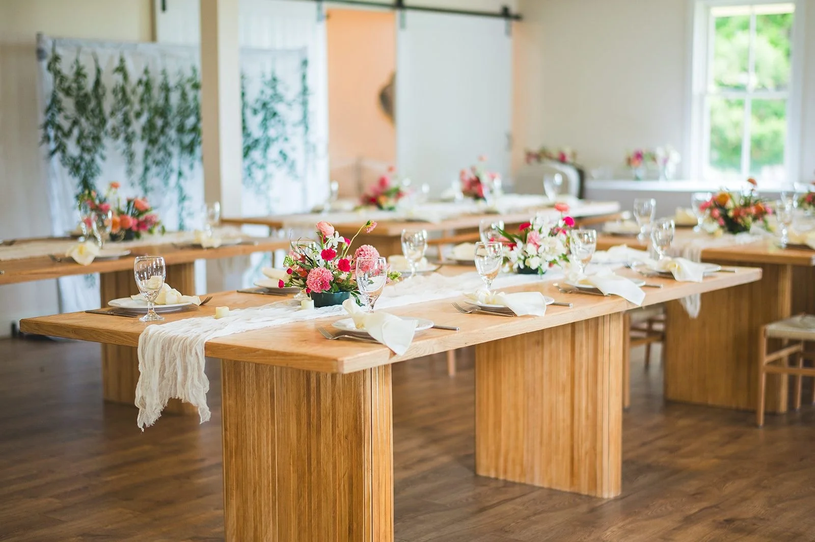 An indoor wedding reception with pink floral centerpieces on white gauze runners topping statement tables crafted from warm-tone wood.