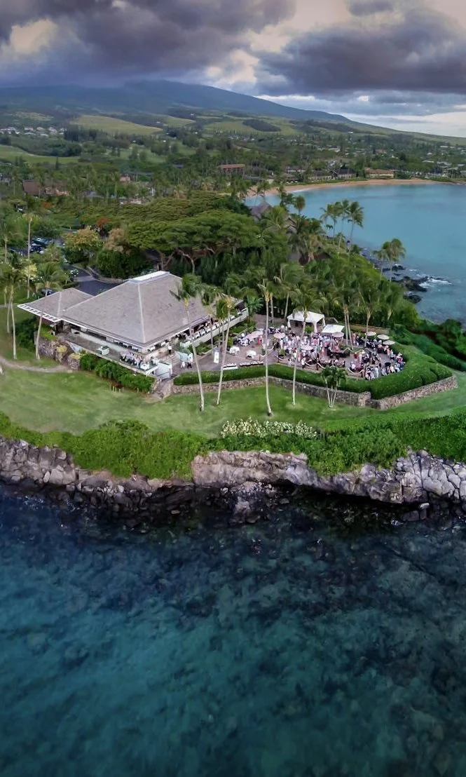 An aerial view of Merriman's restaurant on a grassy cliffside lot over the brilliant blue ocean.