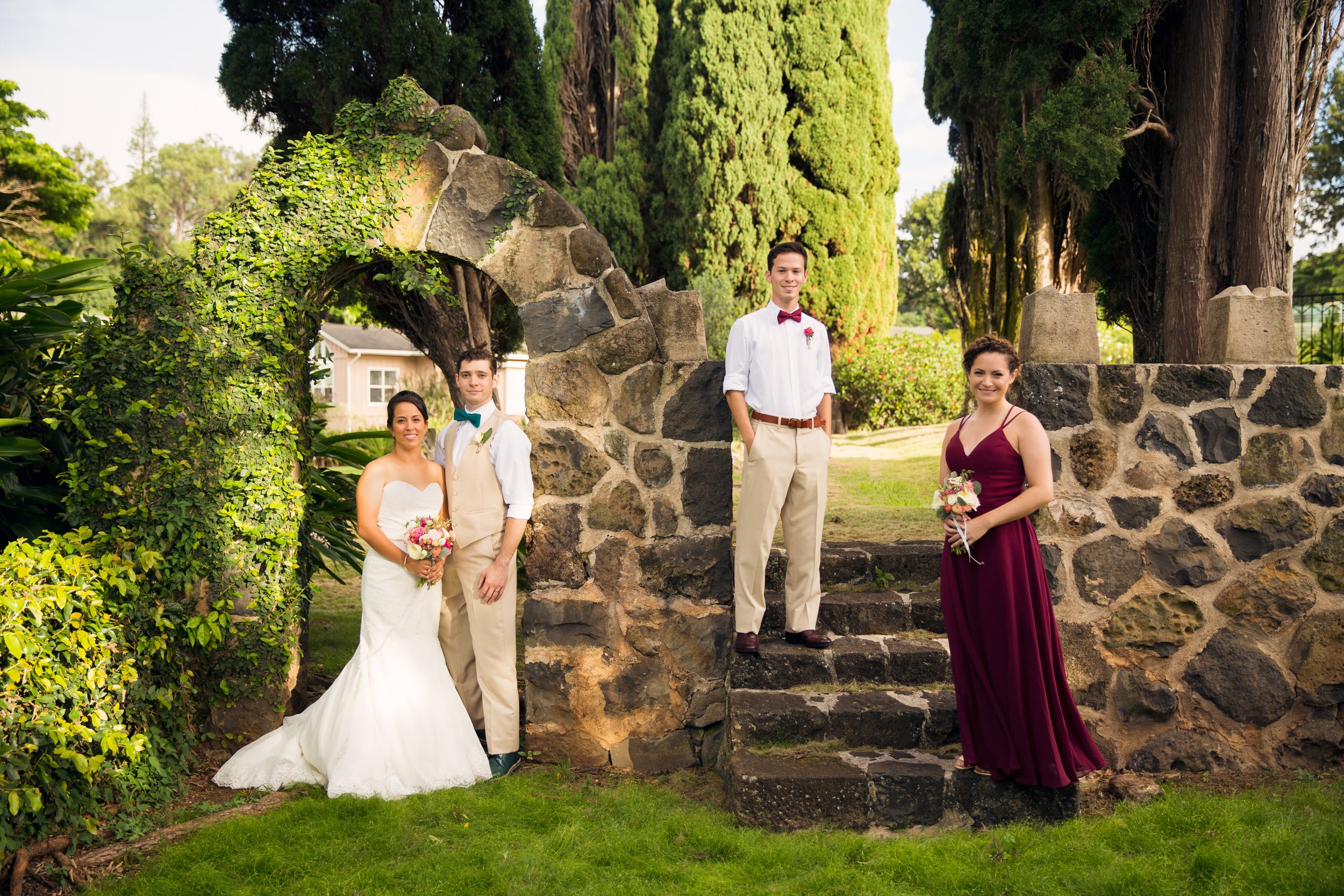 A small wedding party posed outdoors beside a vine-covered stone archway