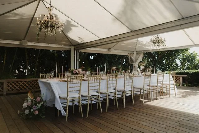 A wedding reception tablescape on a wooden porch beneath a white tent covering.