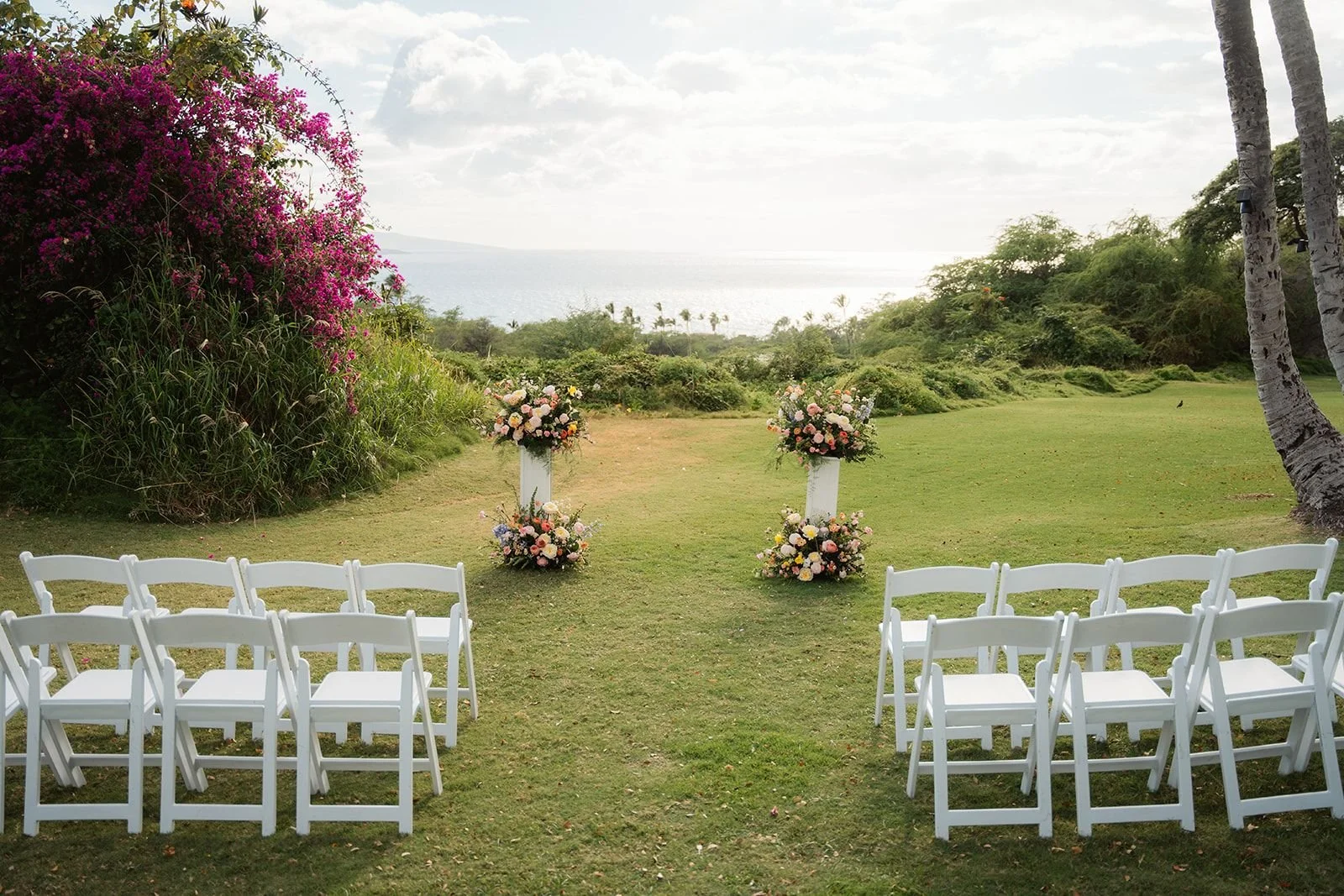 A colorful outdoor Maui wedding with two ceremony pedestals of flowers beside a giant bougainvillea bush before an ocean view.