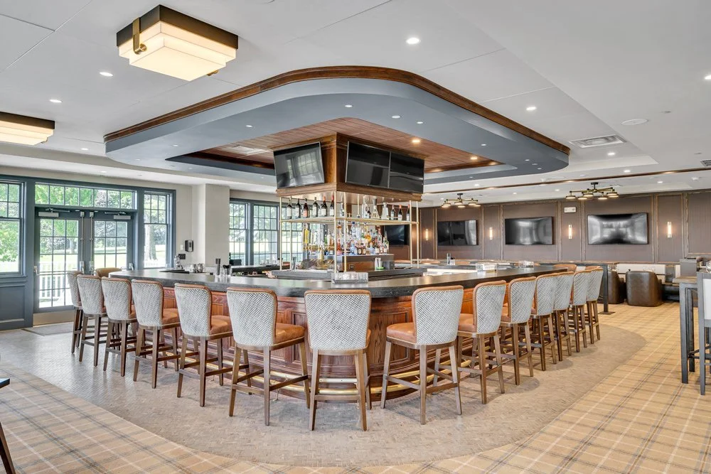 The country club social hub, the bar, with a complete rebuild after a fire. Showcasing its plaid carpet, bright sunlight, and ample dining seating.