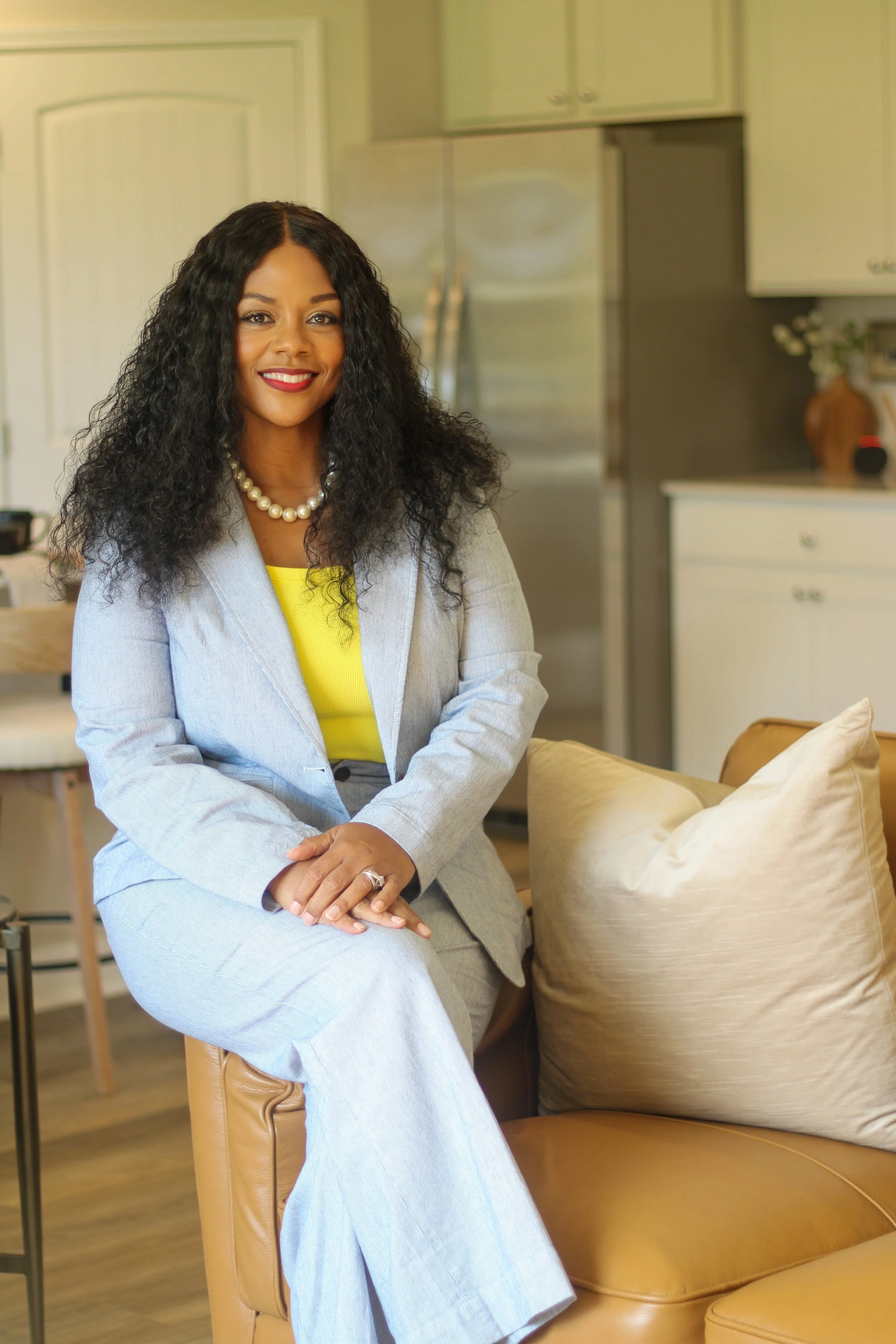 A woman with long curly black hair and a pearl necklace sitting on a tan leather sofa in a modern kitchen.
