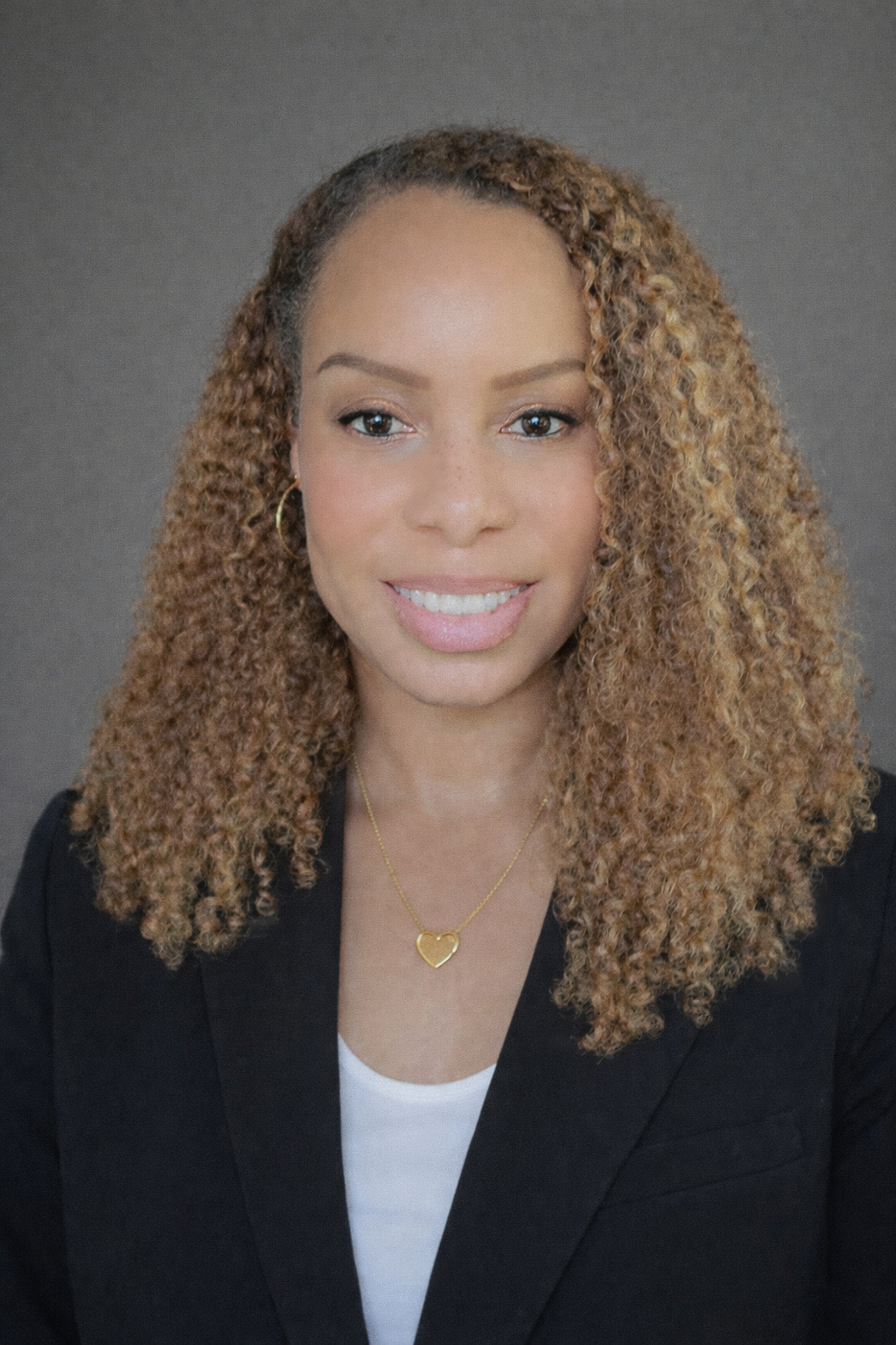Professional headshot of a woman with curly, honey-blonde hair, wearing a black blazer, white top, gold hoop earring, and a gold heart-shaped necklace, smiling against a gray background.