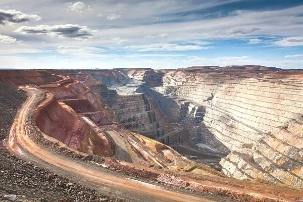 A winding dirt road runs along the edge of a large canyon with layered rock formations, under a partly cloudy sky.
