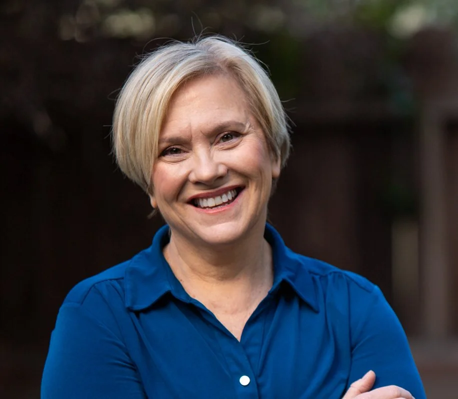 A smiling woman with short blonde hair wearing a blue collared shirt standing outdoors, with a blurred background of trees and a fence.
