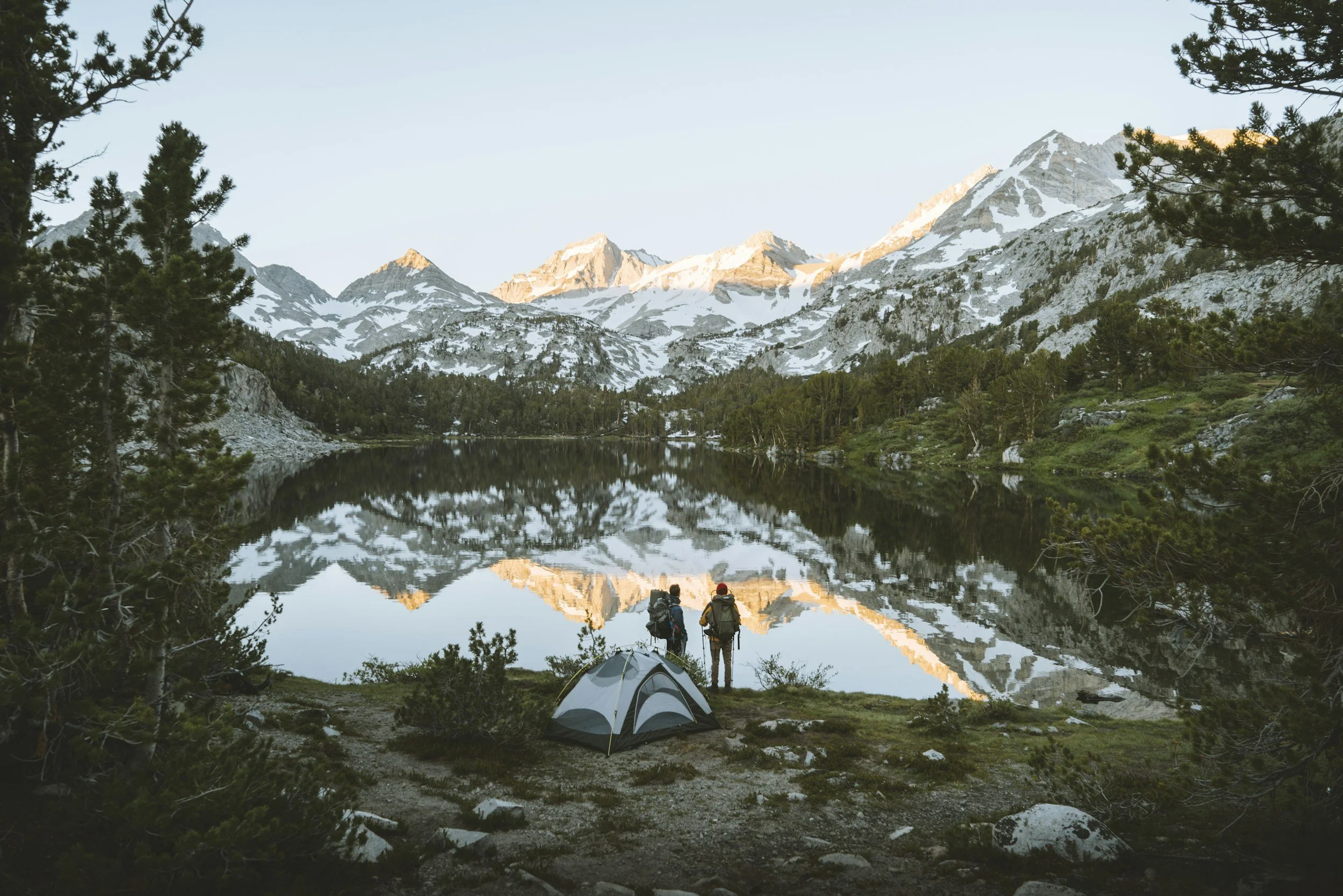 Two hikers with backpacks standing by a lake surrounded by pine trees, with snow-capped mountains in the background during sunset.