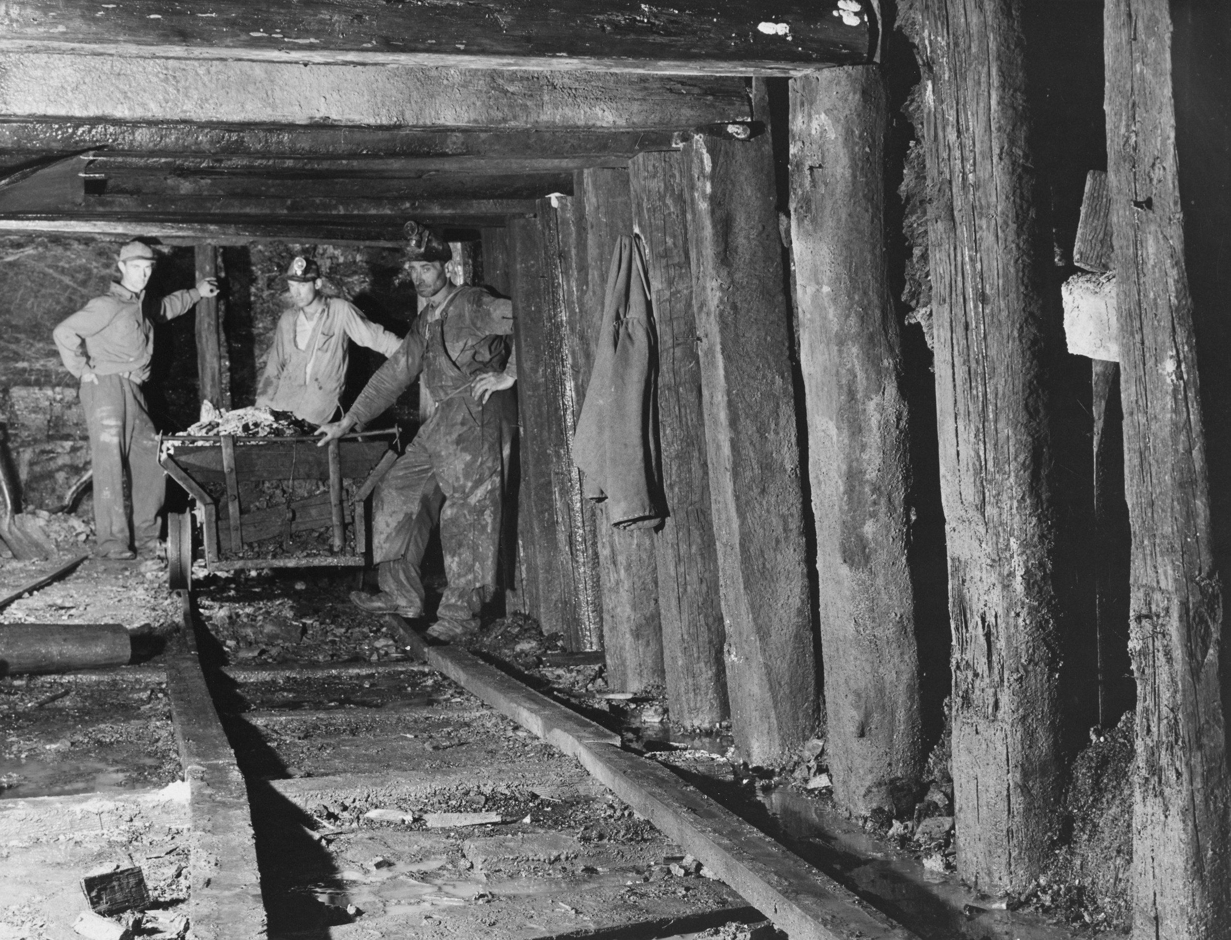 Three workers, dressed in work clothes and helmets, standing inside a mine with wooden supports and a cart loaded with ore.