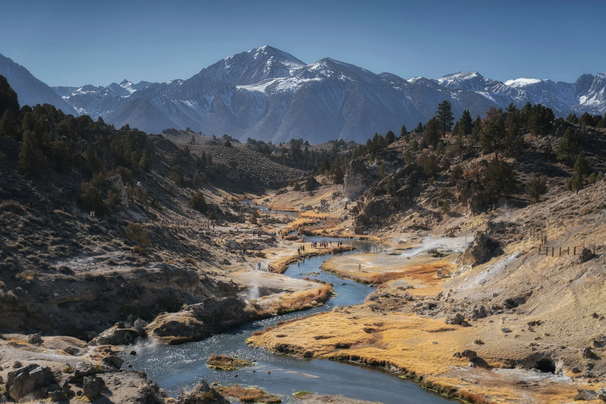Scenic mountain landscape with snow-capped peaks, a winding river, and a trail with hikers in the foreground.