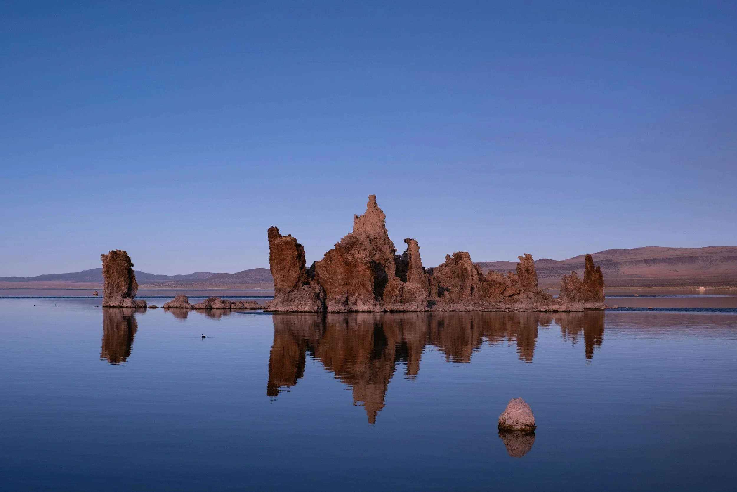 Large rock formations in a lake with clear reflections, distant mountains, and a blue sky.