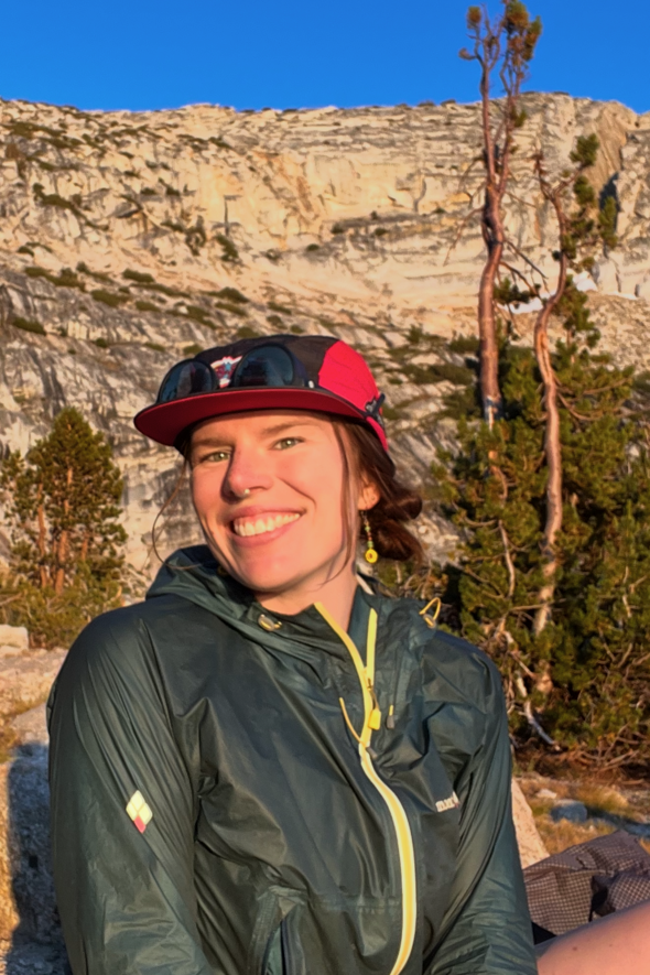 A woman with a reddish-brown braid wearing a black and red cap, sunglasses on top, and a dark green outdoor jacket, smiling at the camera outdoors with trees and a rocky cliff in the background during sunset.