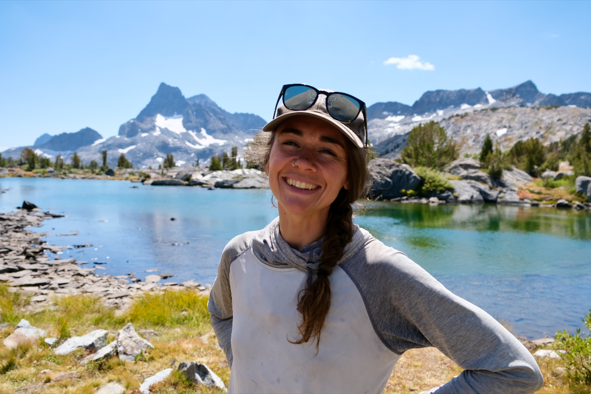 Young woman smiling outdoors near a mountain lake with snow-capped peaks in the background.