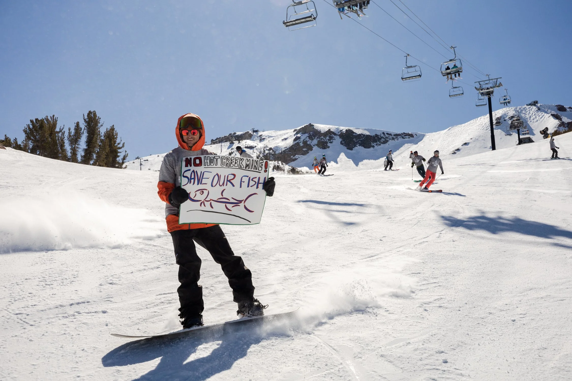A person snowboarding on a snowy mountain, holding a protest sign that reads, 'NO HOT CREEK DRAIN! SAVE OUR FISH', with ski lifts and other skiers in the background under a clear blue sky.