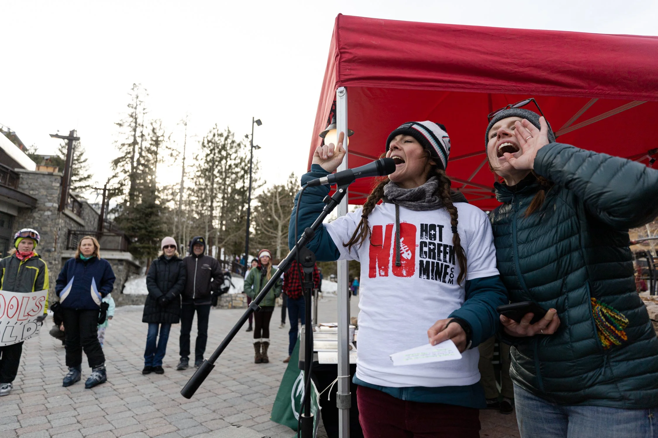 A woman speaking into a microphone at a rally or protest, wearing a white t-shirt with red and black text, a gray scarf, and a winter hat with braids. She is holding a piece of paper. Beside her, a man is leaning in, cupping his hand around his mouth to amplify his voice or shout. In the background, several people are standing outdoors under a cloudy sky, with some holding signs, and a red canopy covers part of the scene.