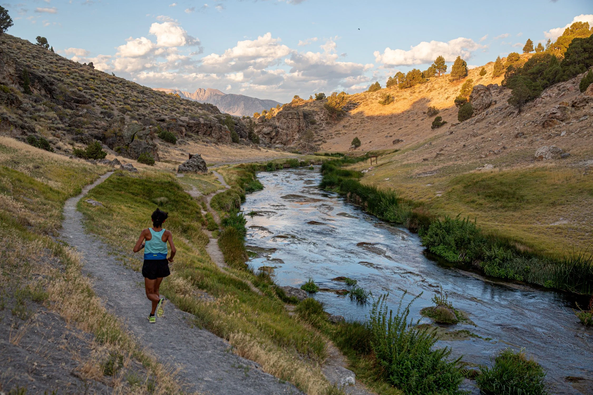 A person jogging on a dirt trail beside a river in a mountainous landscape with scattered trees and a partly cloudy sky.