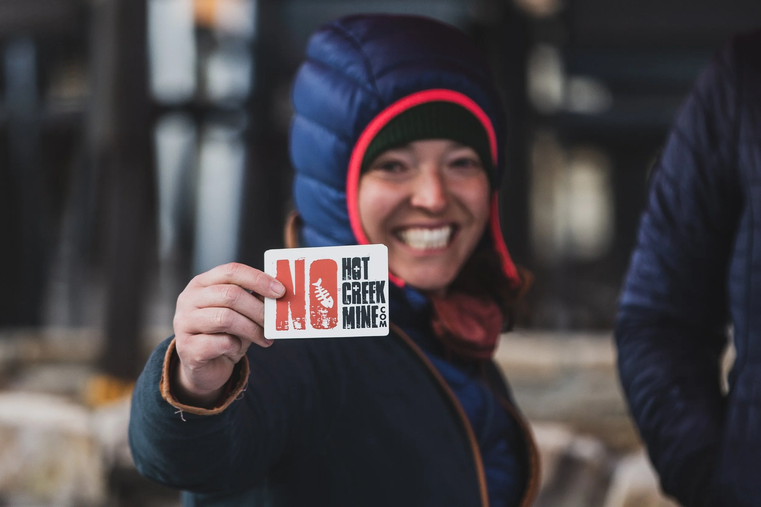 Woman smiling and holding a sticker that reads 'NO HOT CREEK MINES' in front of her face, wearing a winter jacket and a puffy hat.