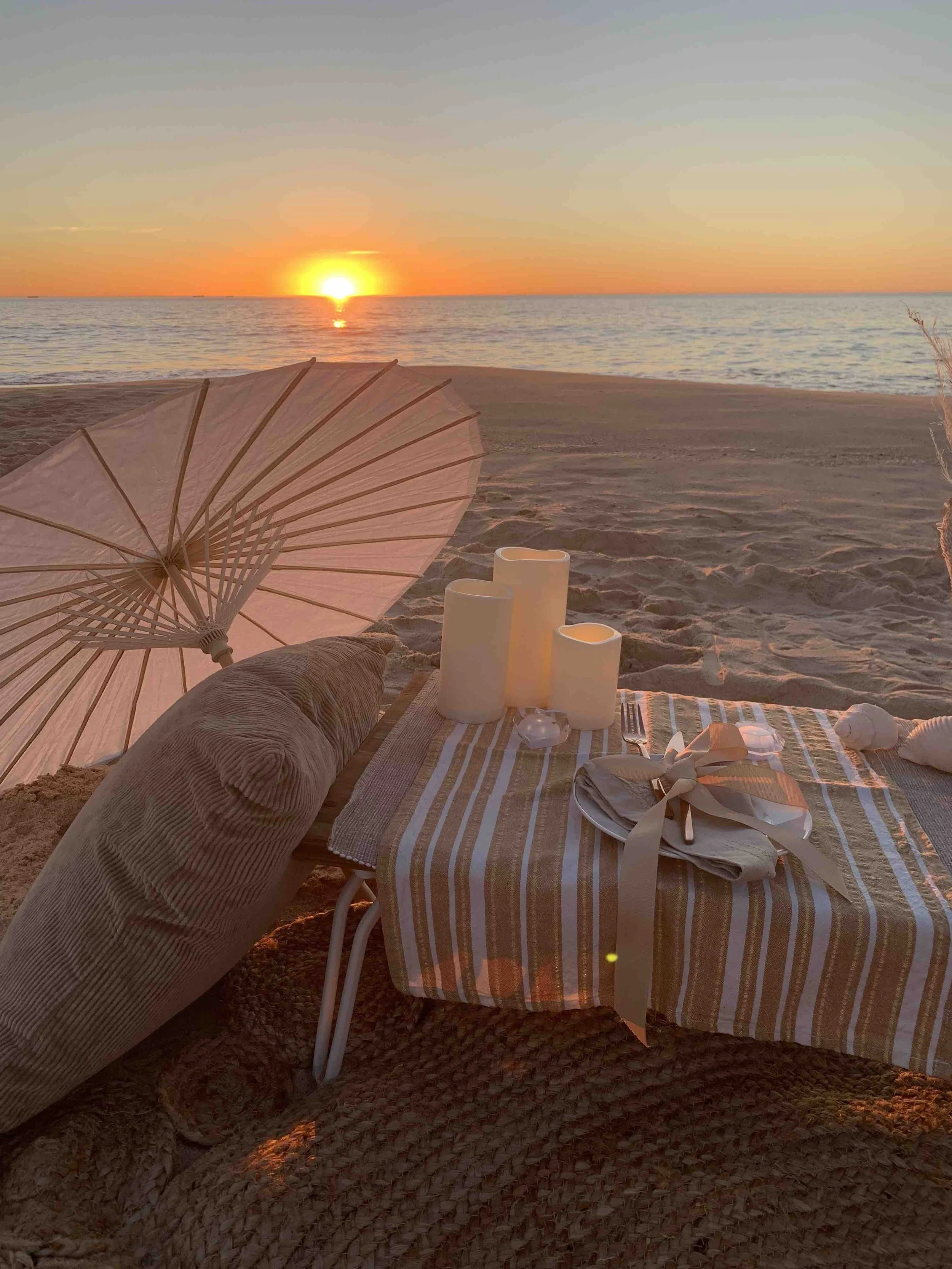 Candles and cutlery sit on a table on sand with the sun setting