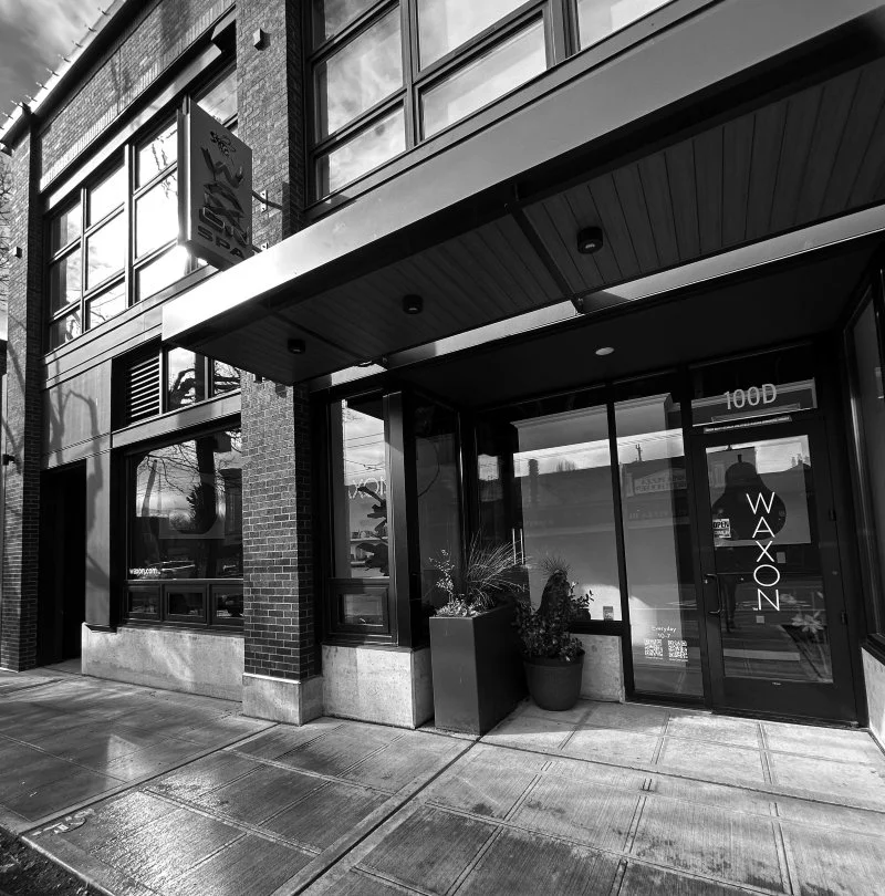 Black and white photo of the storefront for Waxon, a business located at 100D, with large glass windows and a glass door, two potted plants outside, and an awning above the entrance.