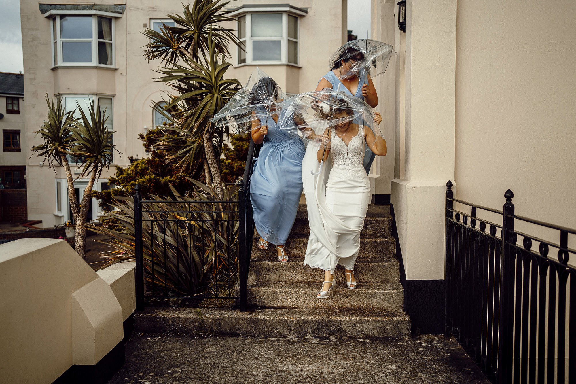 Bride and bridesmaids with clear umbrellas descending steps on a windy day, wearing wedding and blue dresses, near a building with palm trees.