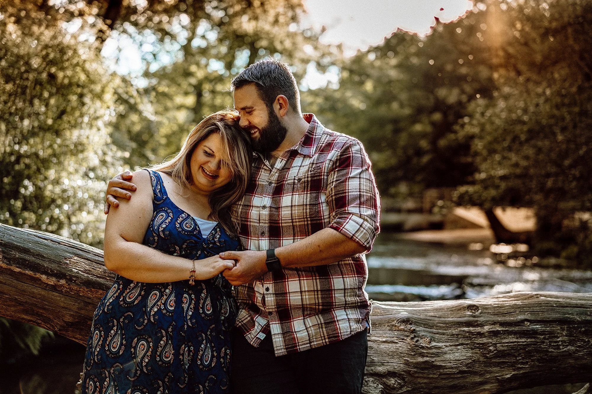 Couple embracing outdoors near a log, surrounded by trees and sunlight.