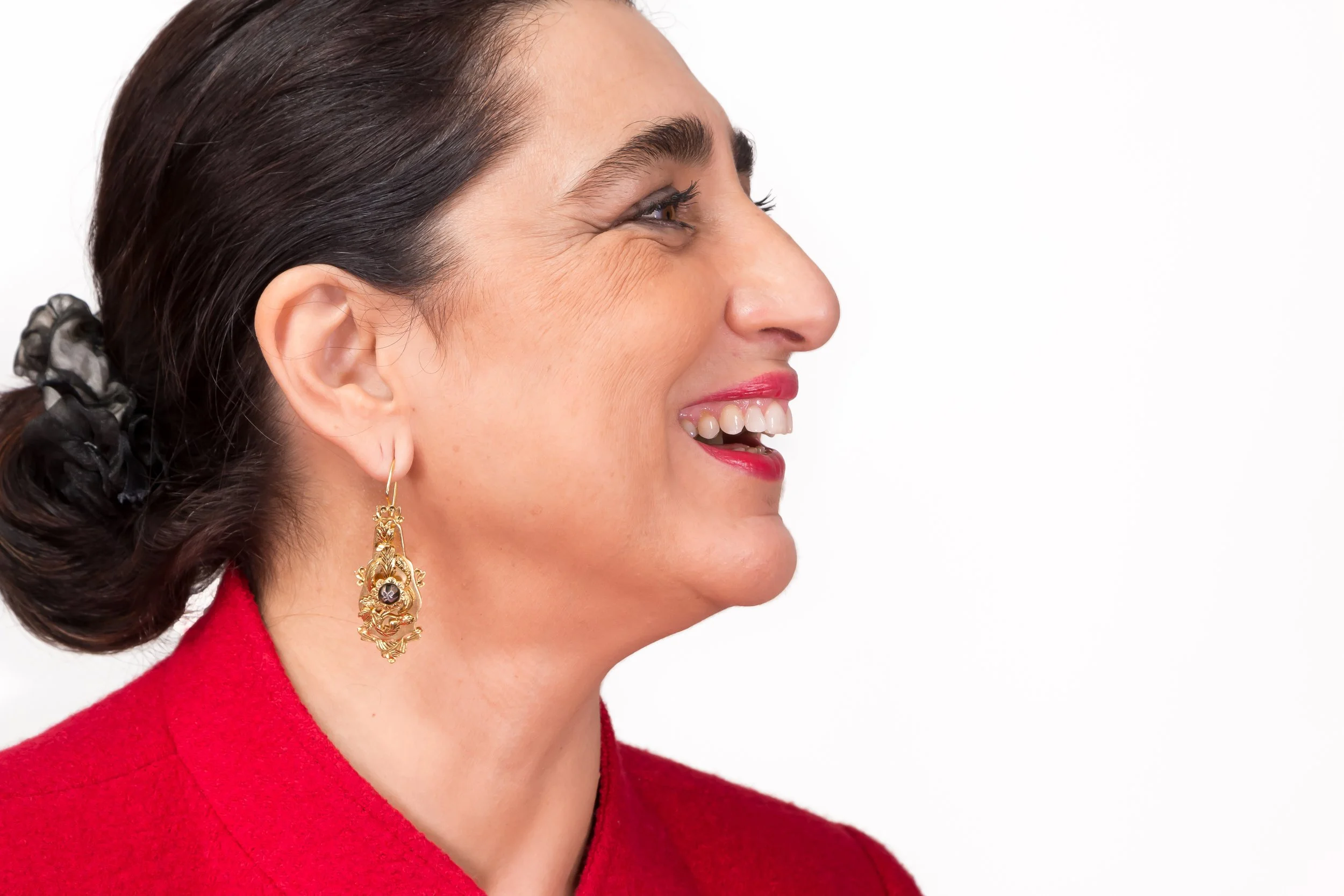 Mulher sorrindo de perfil, com cabelo escuro preso com lenço, usando brincos dourados e roupa vermelha.