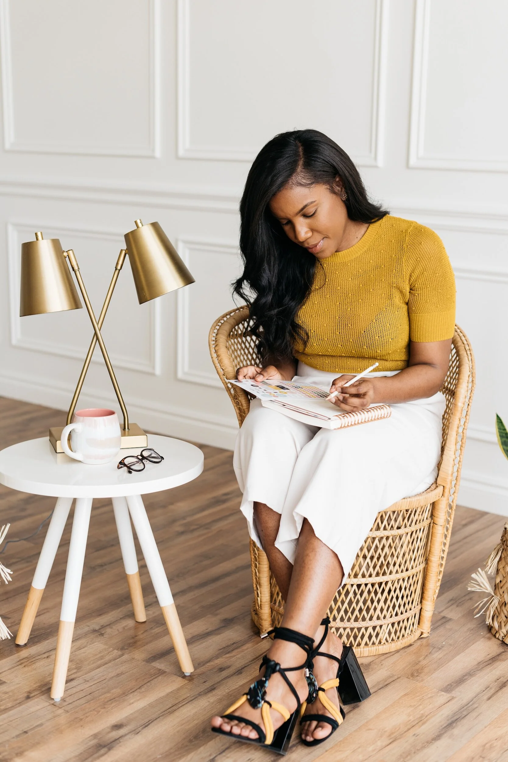 A woman with long black hair, wearing a yellow short-sleeve sweater and white wide-leg pants, sitting in a wicker chair, writing in a notebook.
