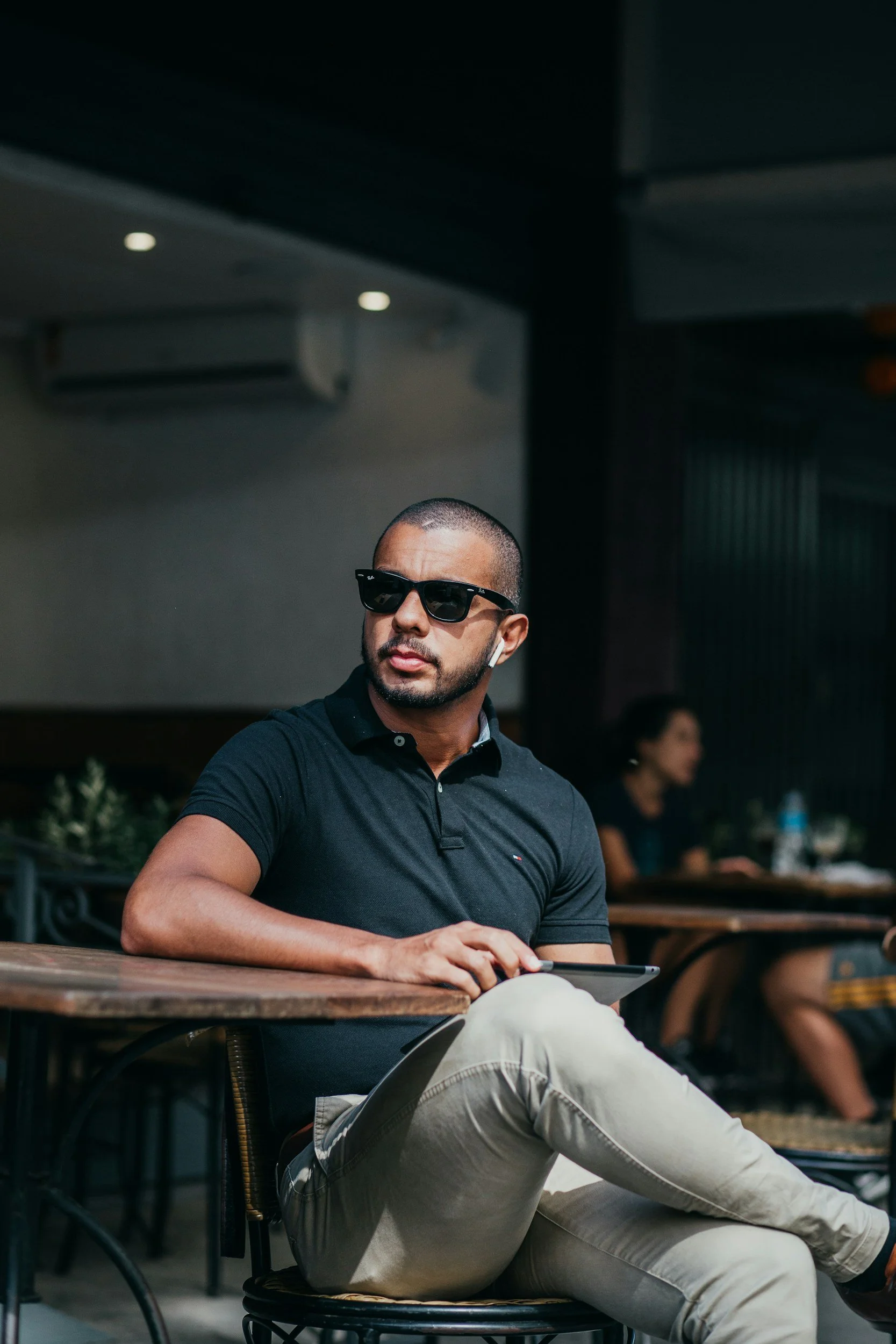 A man wearing sunglasses and AirPods sitting at an outdoor café table, holding a tablet with other people visible in the background.