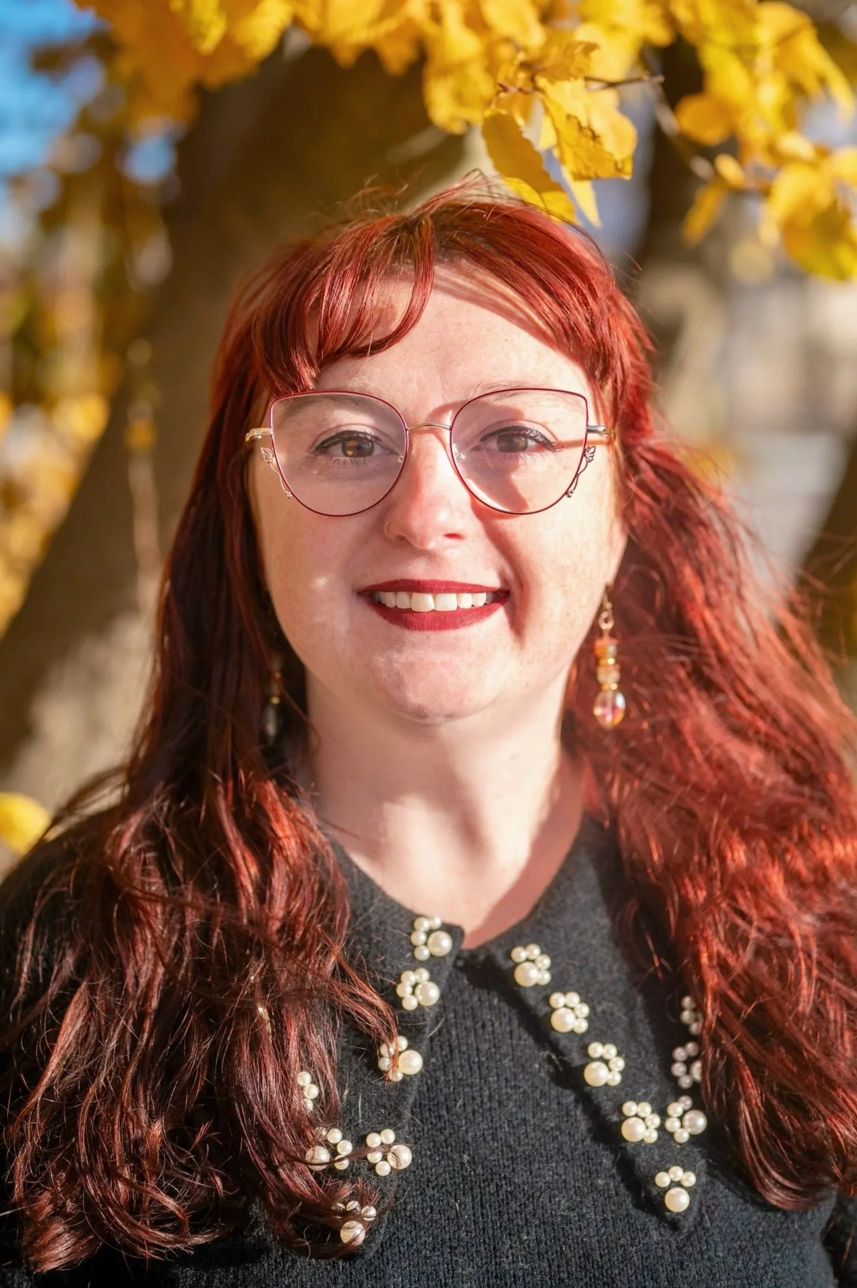 A female therapist with red hair and glasses smiling outdoors in front of a tree with yellow leaves.