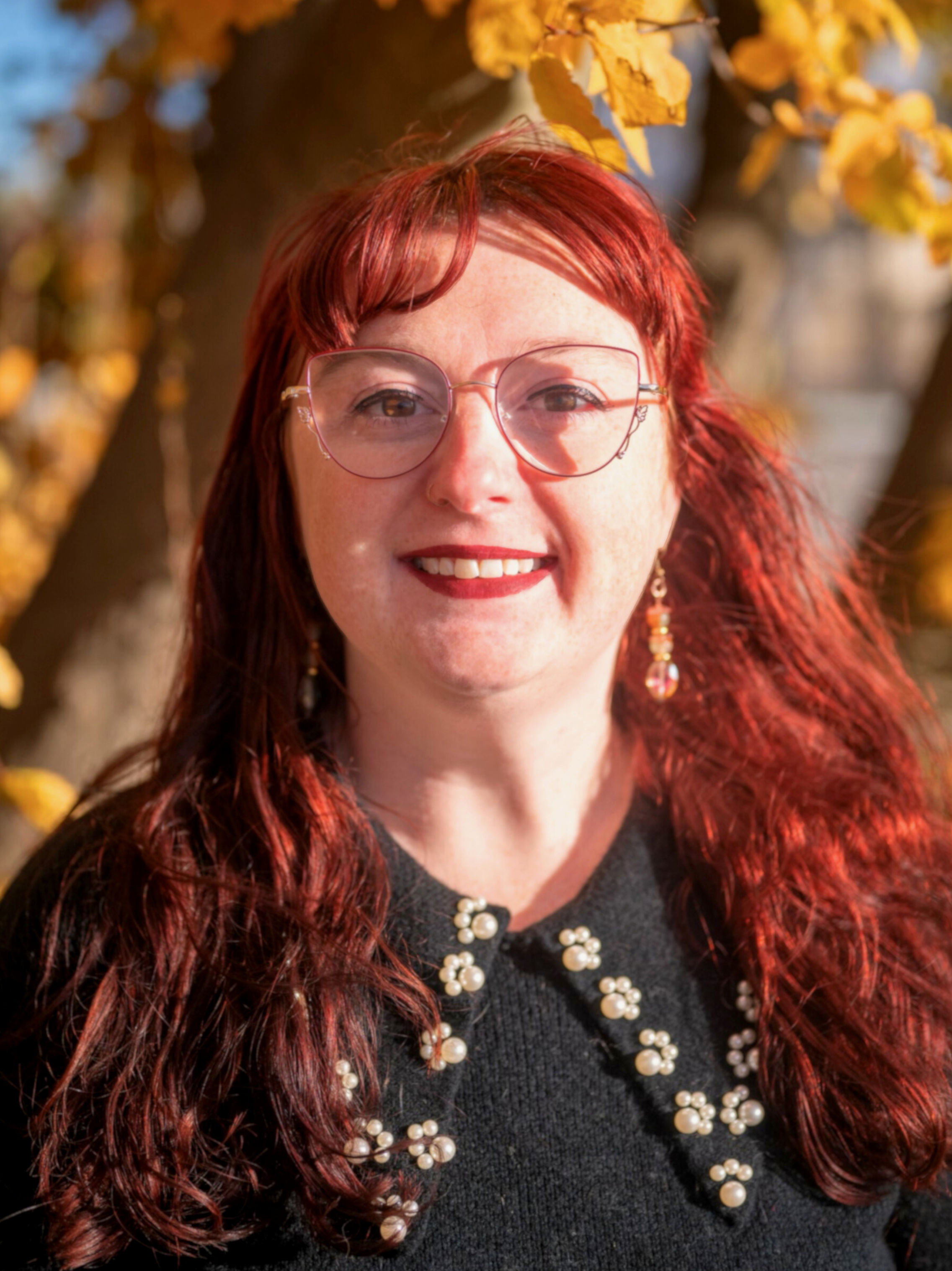 A female therapist with red hair, glasses, and earrings, smiling outdoors with autumn leaves in the background.