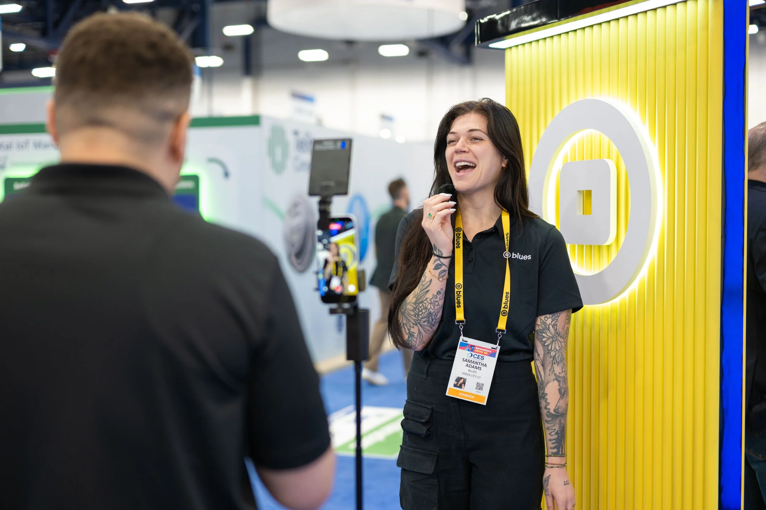 A woman with tattoos on her arms and long dark hair wearing a black shirt with a lanyard and name badge, smiling and talking to a man in a black shirt at a conference booth with a large yellow and white logo in the background.
