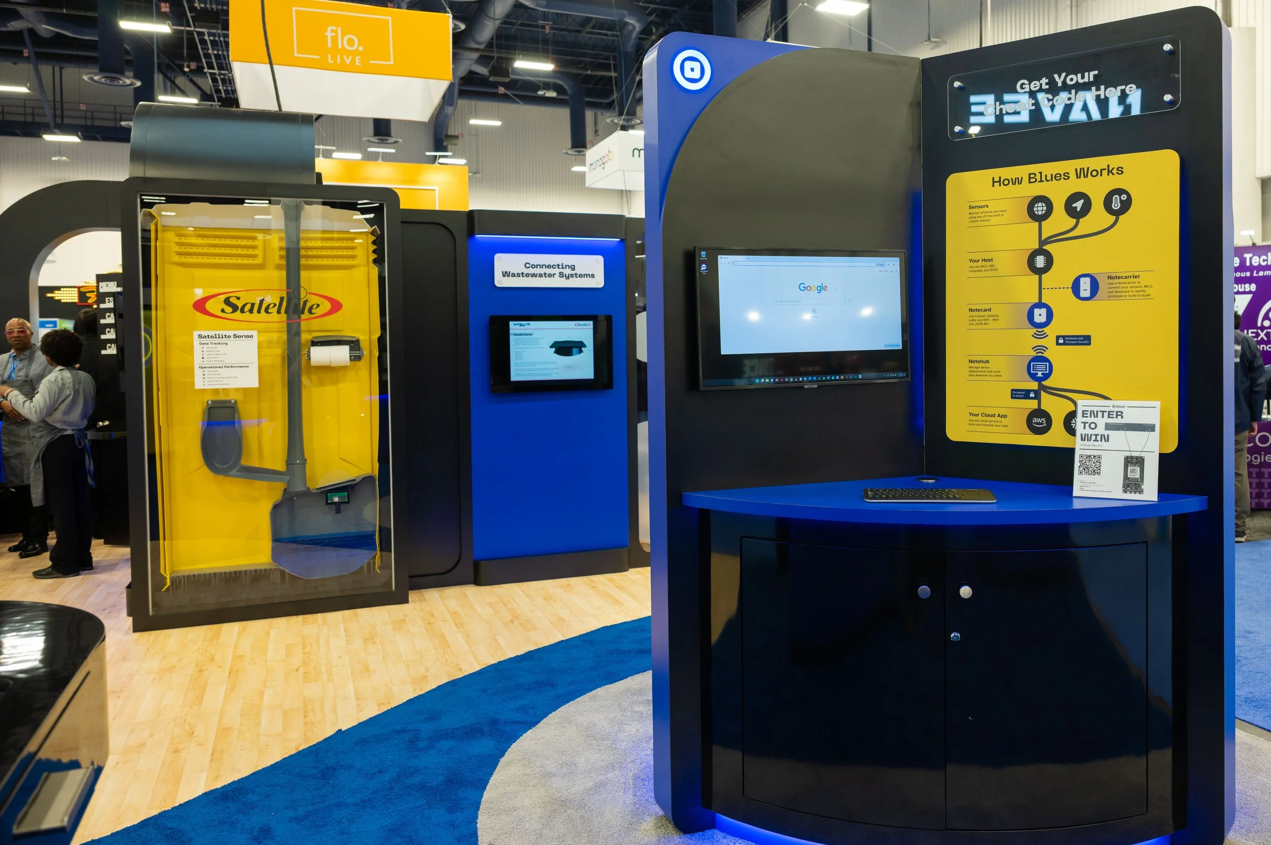 Exhibit at a technology conference showing a yellow satellite hardware box, a blue wastewater system display, and an interactive station with a monitor and informational signs about how blues works.