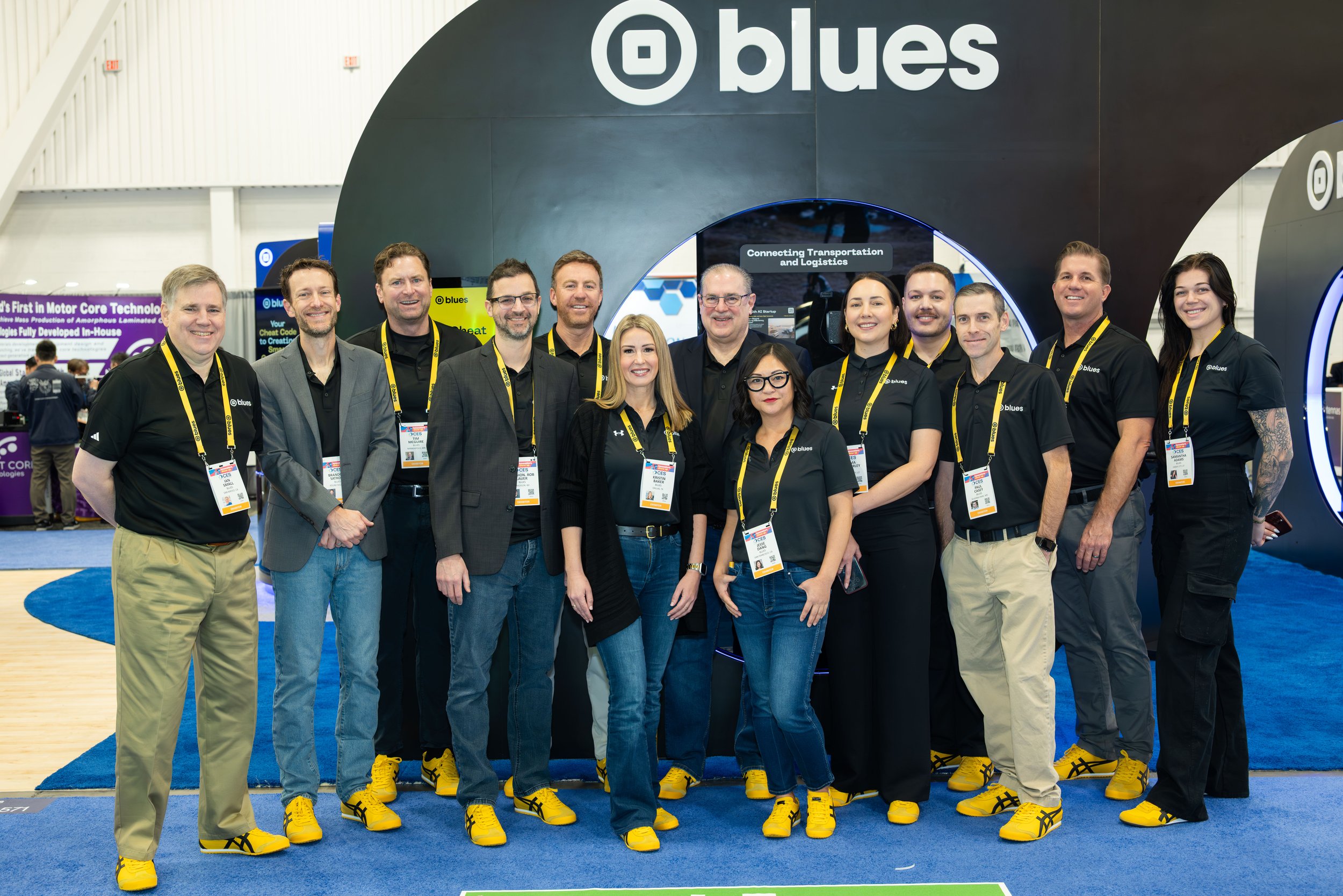A group of eleven people, both men and women, standing together at a trade show or conference in front of a large black booth with the Instagram logo and the word 'blues'. They are all wearing black or gray shirts, with some in blazers, and yellow sn