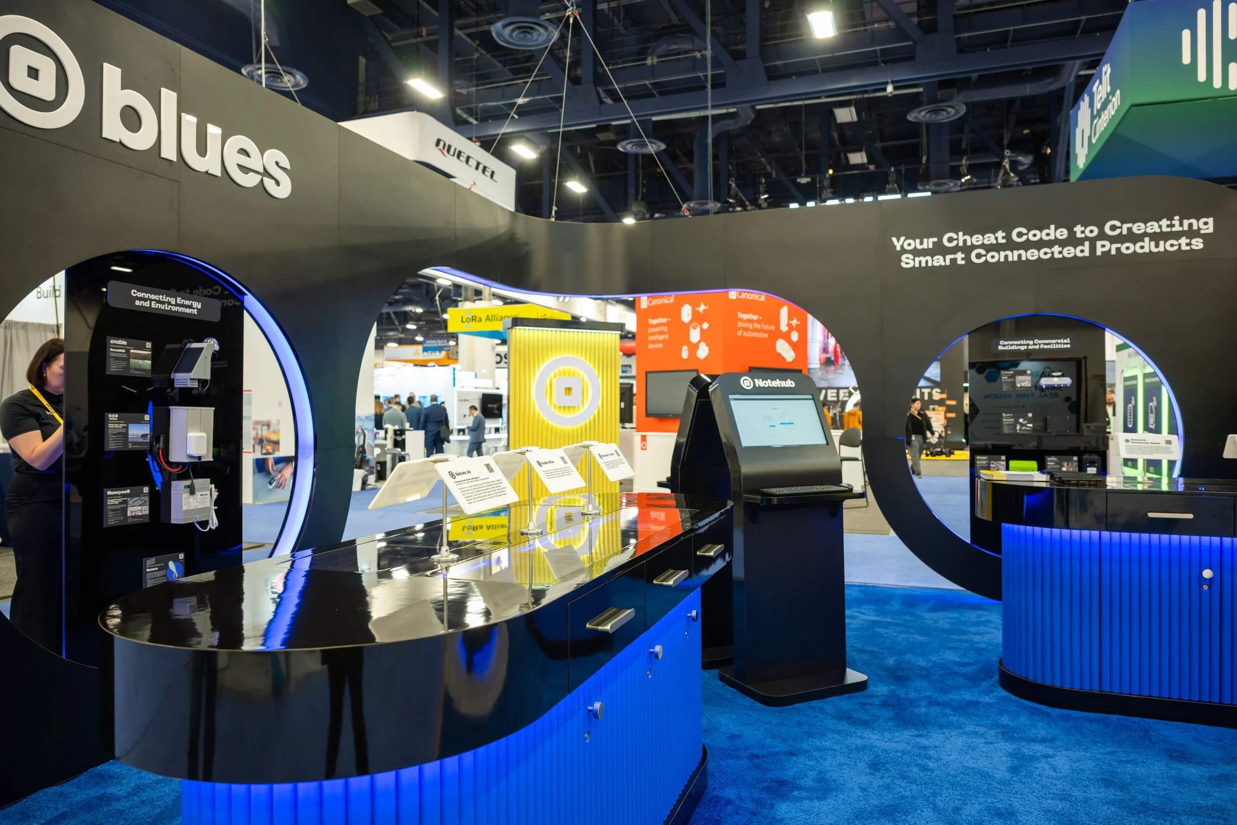 Exhibit booth at a tech trade show featuring smart connected products, including energy and environment devices, with signage reading 'Your Cheat Code to Creating Smart Connected Products'.