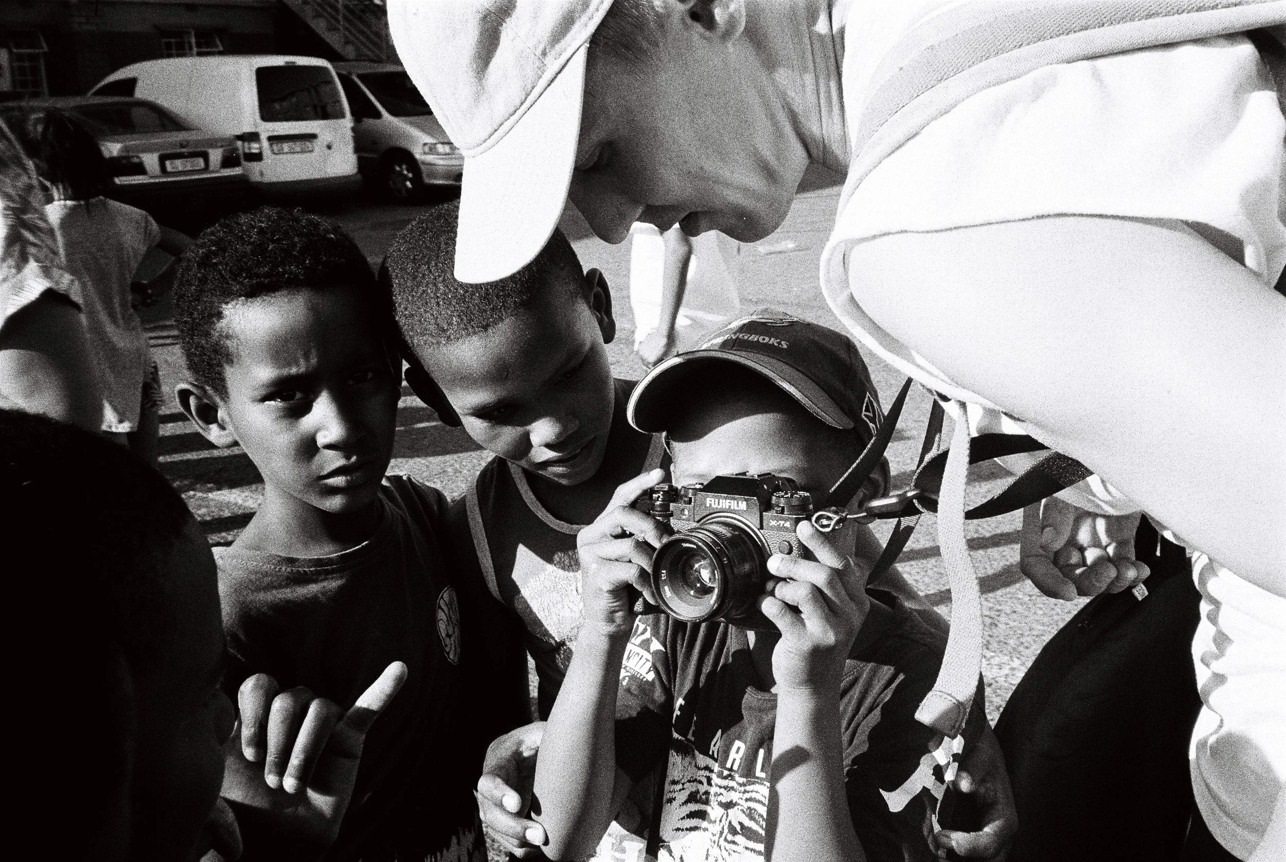 A group of children gathered closely around a person taking a photo with a Fujifilm camera in an outdoor setting with parked cars in the background.