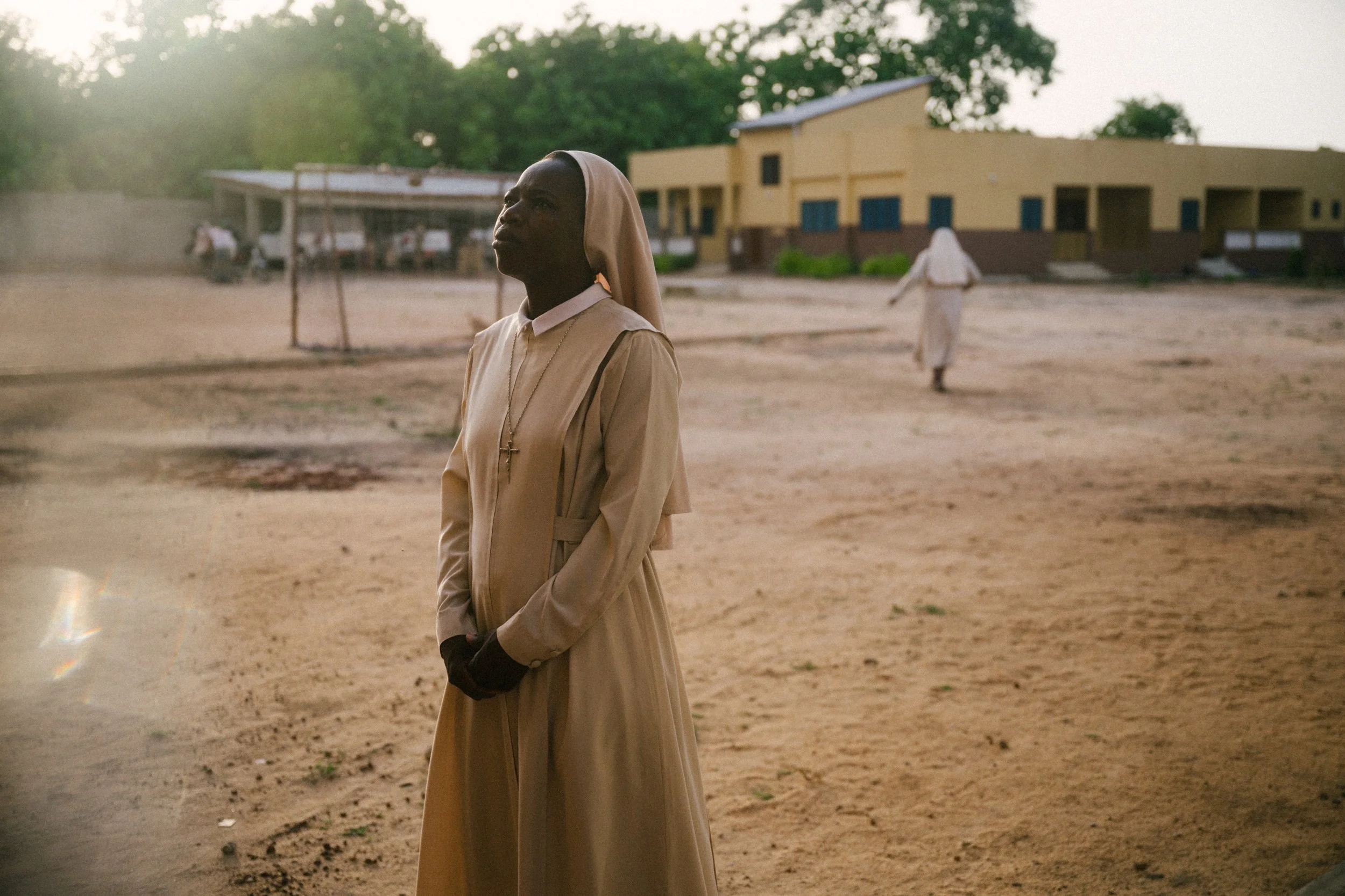 A nun dressed in beige robes with a cross necklace standing outside on dusty ground, with a large building in the background and another nun walking away.