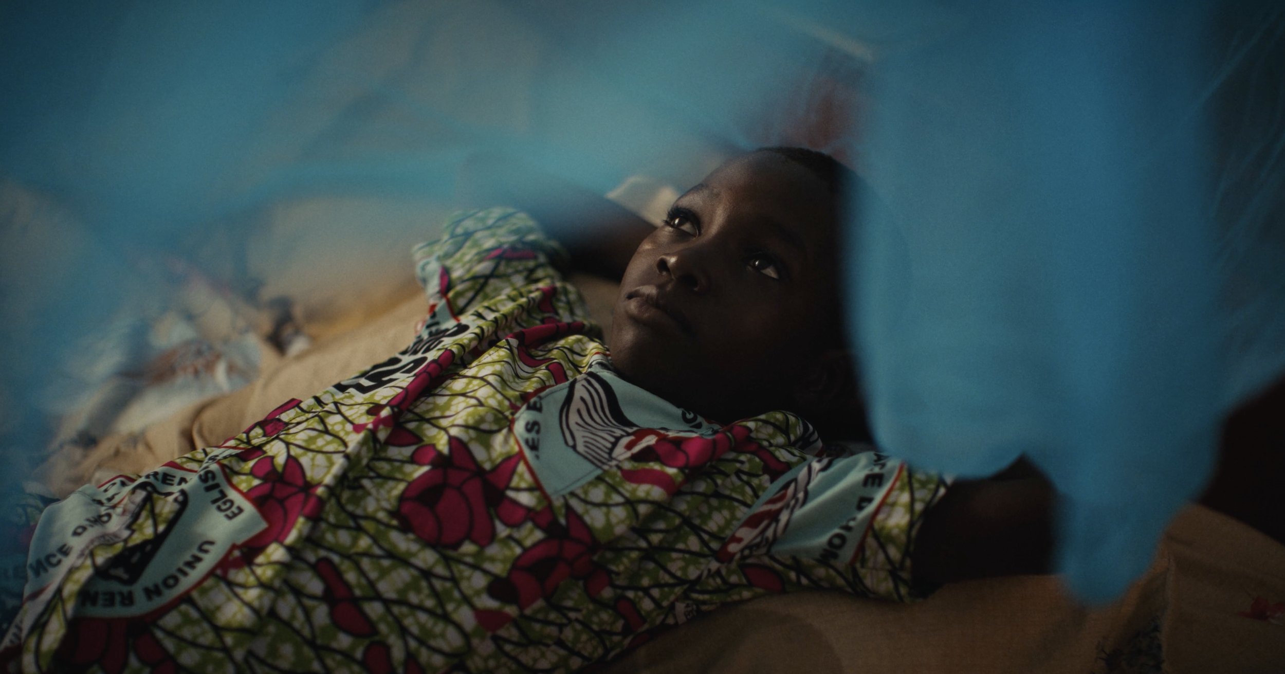 A young girl lying in a hospital bed, looking up thoughtfully, with IV line attached to her arm and a blue medical curtain partially obscuring the view.