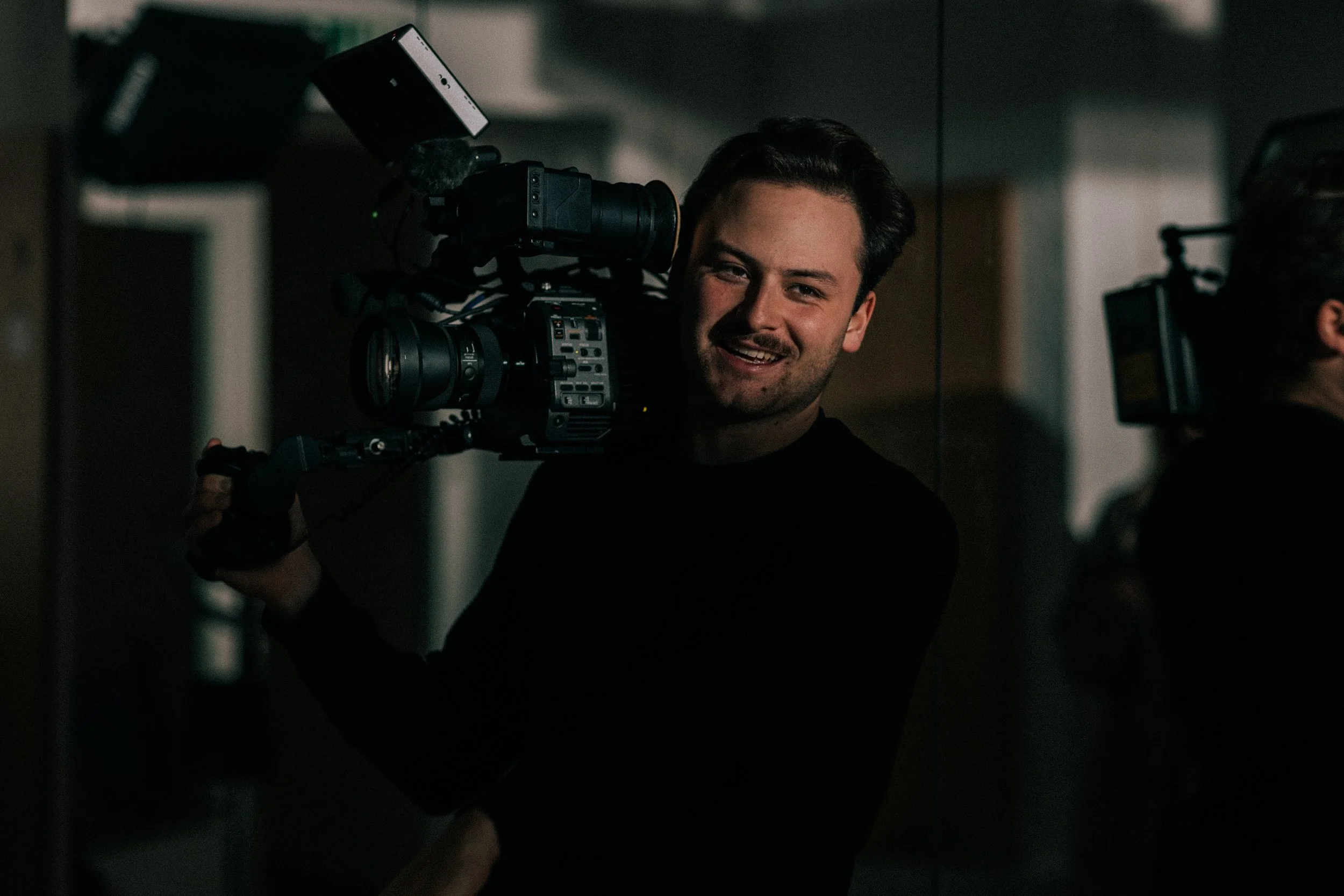 A man smiling while holding a professional video camera on his shoulder, in a dark indoor setting.