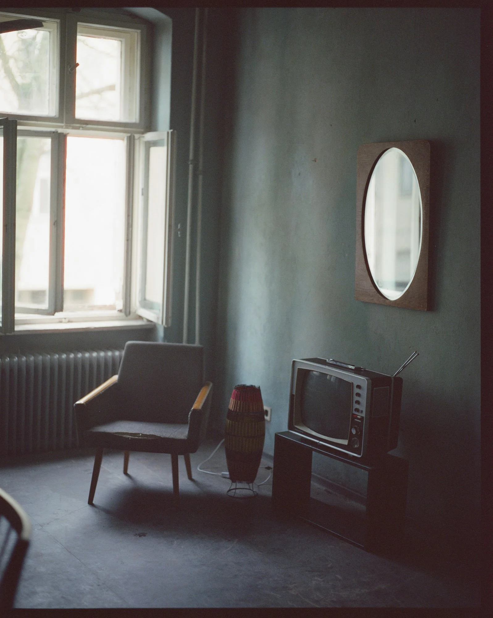 A vintage TV on a stand, a black armchair with wooden arms, a tall colorful woven floor lamp, an oval mirror on the wall, and an open window with natural light in a room with a dark floor and gray walls.