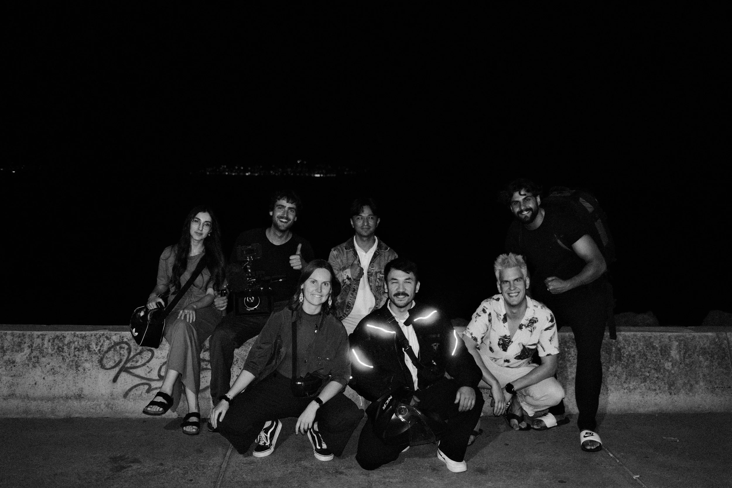 Group of eight young adults smiling at night by a water body with city lights in the background, some people making thumbs up gestures, one person holding a camera, all dressed casually.