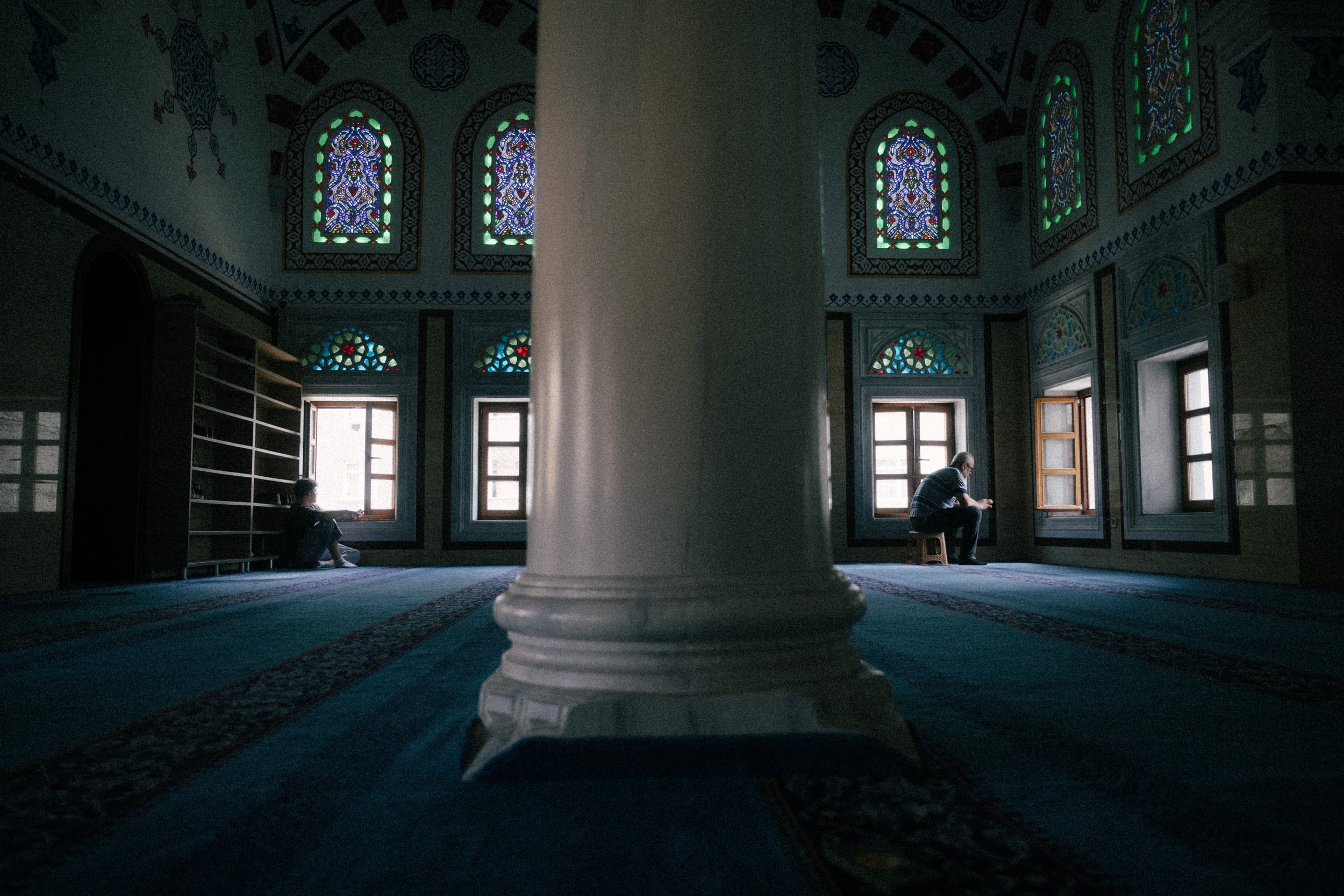 A large prayer room with stained glass windows and patterned walls, divided by a central white column. Two individuals sit silently by the windows, one on the left and one on a small stool on the right.