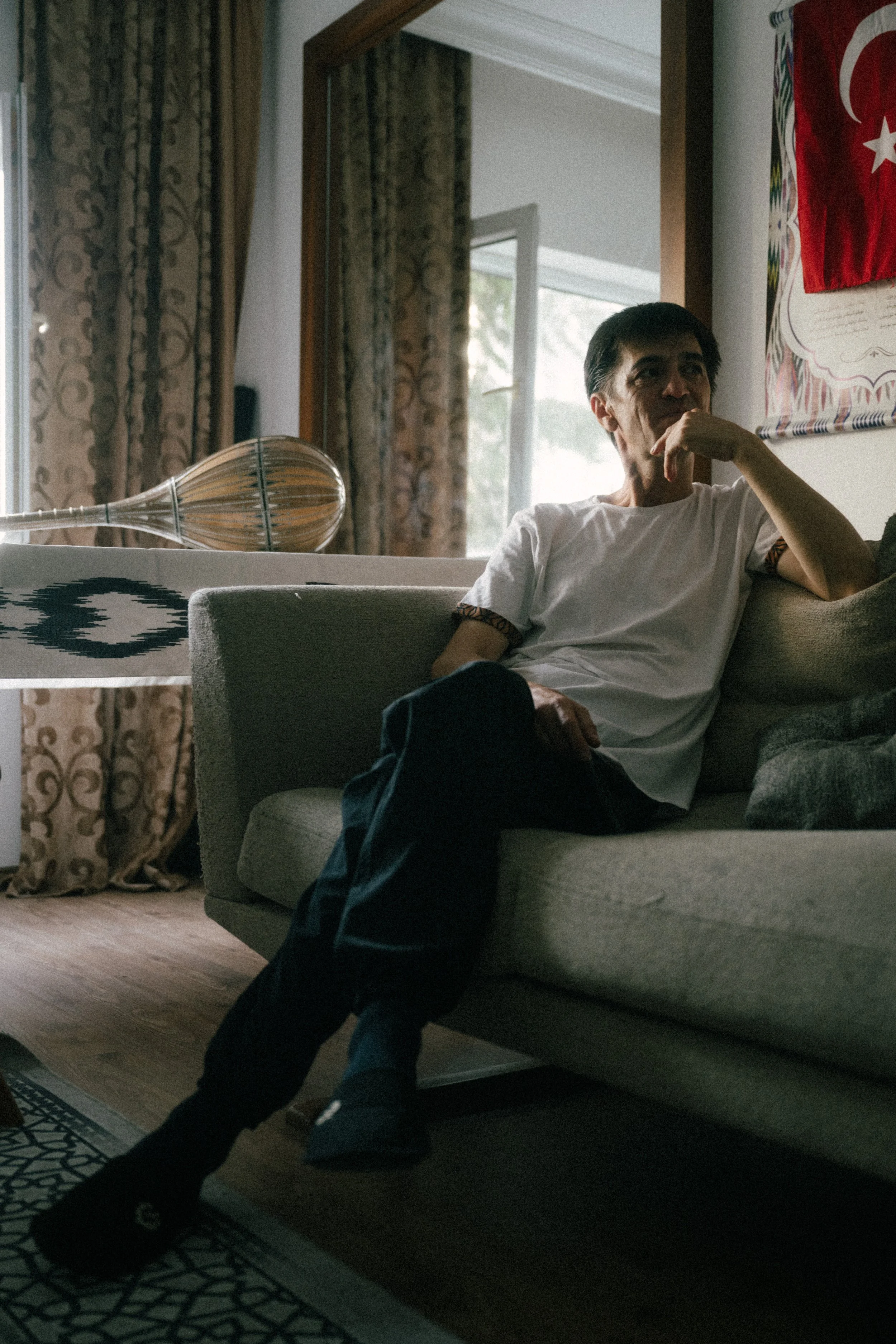Man sitting on a sofa in a room with patterned curtains, Turkish flag artwork, and a glass table with a lamp.