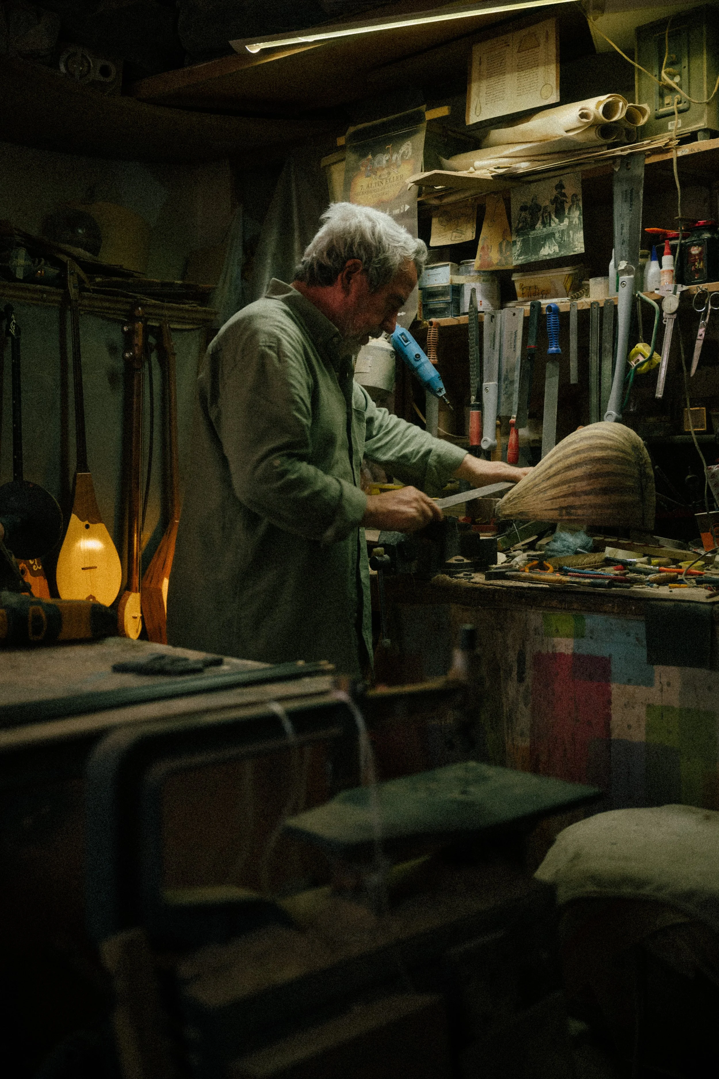 An older man working in a woodworking shop, using a saw on a piece of wood, surrounded by tools, books, and musical instruments in a cluttered workshop.