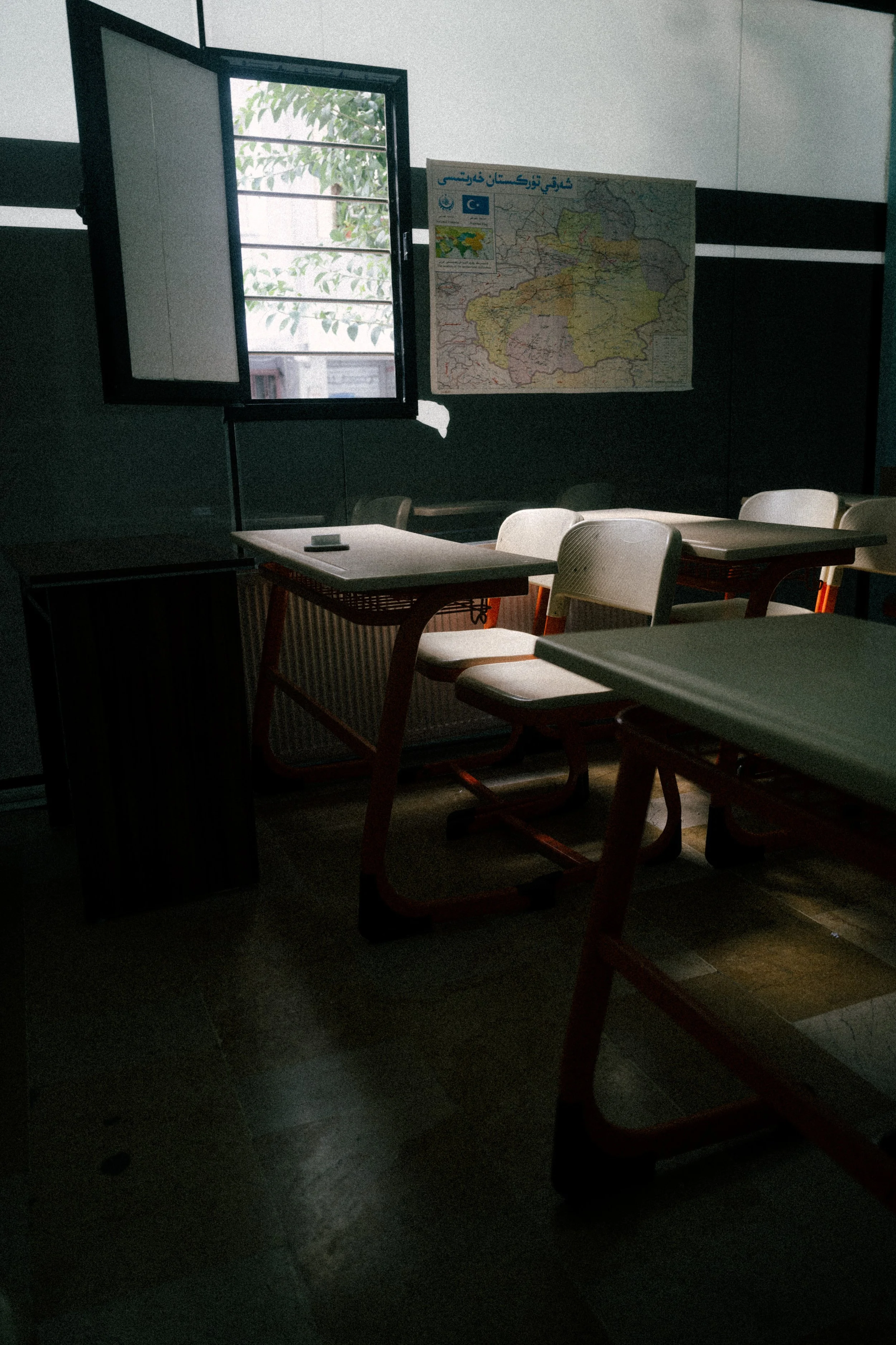 A classroom with open window letting in light, white desks and chairs, a map on the wall, and a radiator under the window.