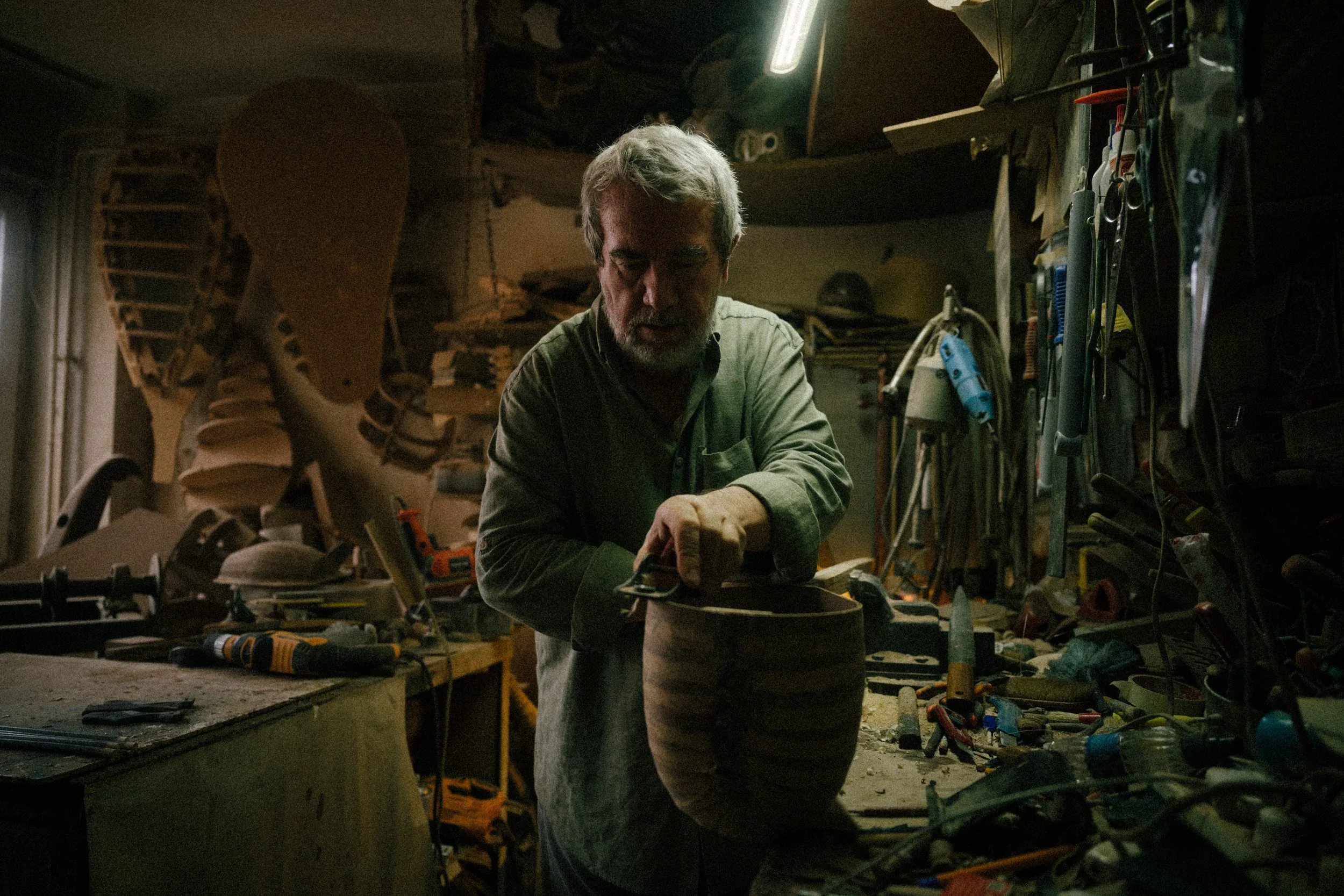 An older man with gray hair and a beard working in a cluttered woodworking workshop, shaping a piece of wood.