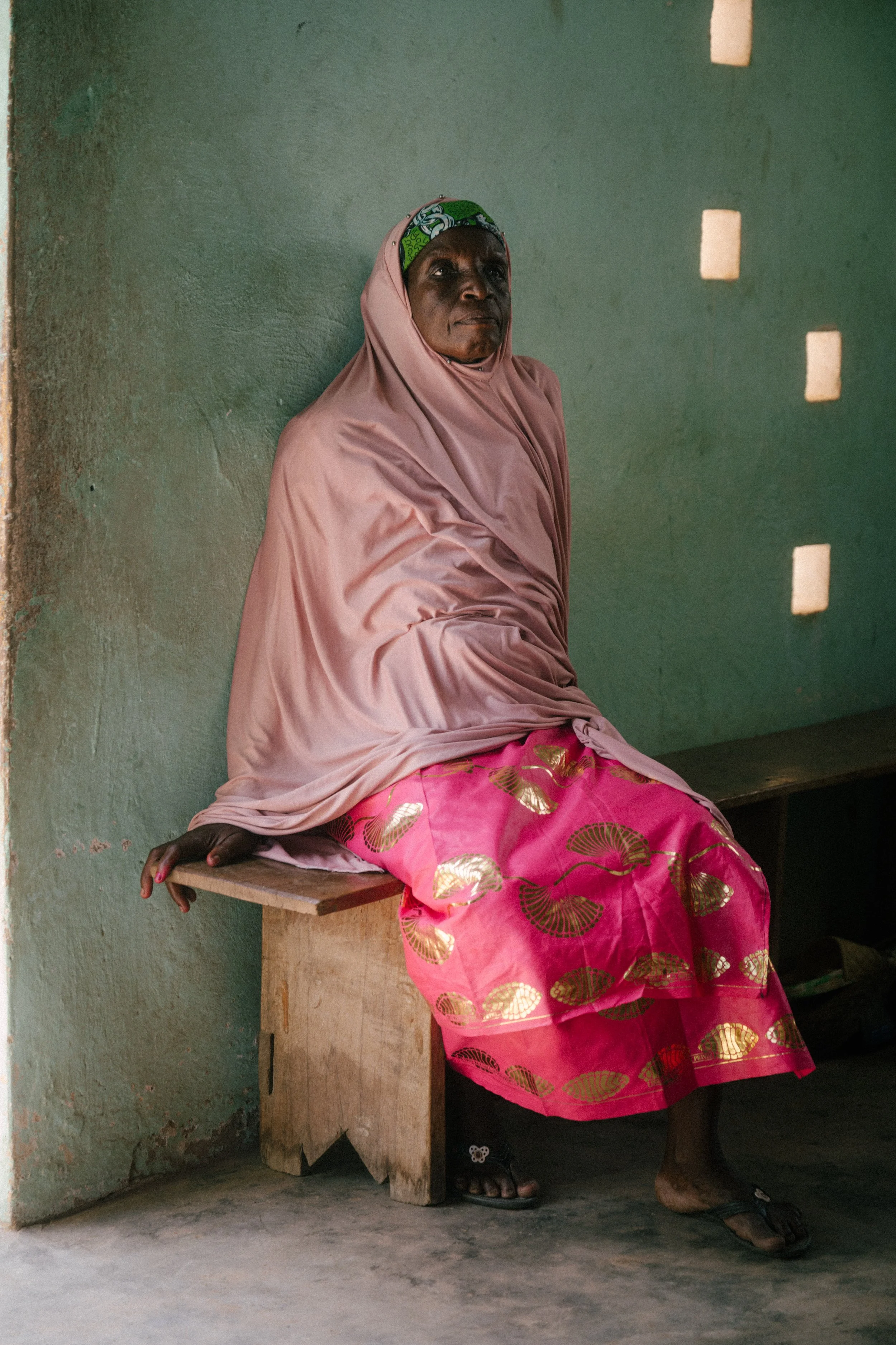 An older woman wearing a pink hijab, pink dress with gold patterns, and a green headscarf sitting on a wooden bench against a green wall indoors.